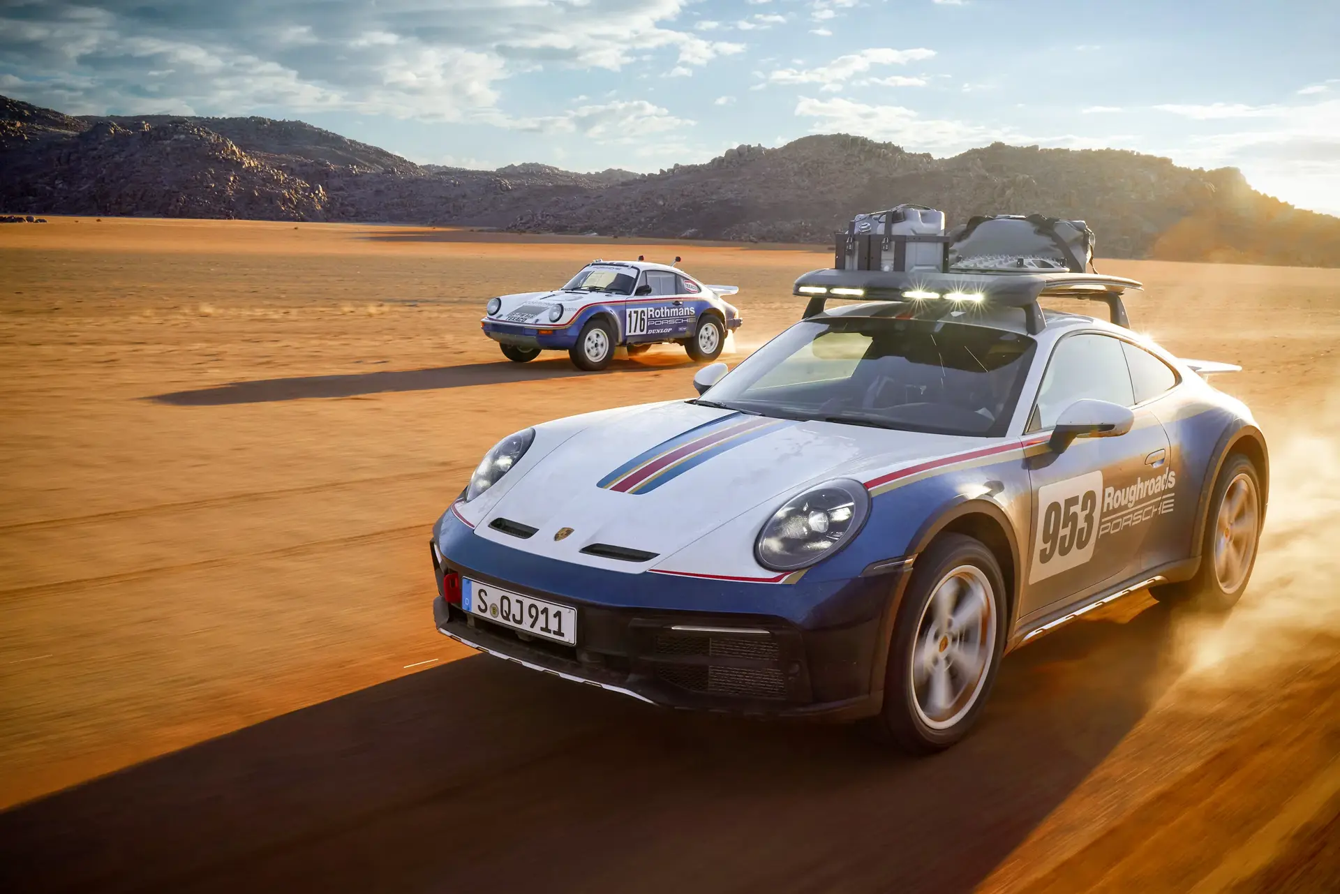 To rallyinspirerede Porsche-sportsvogne kører i høj fart gennem et solbeskinnet ørkenlandskab.