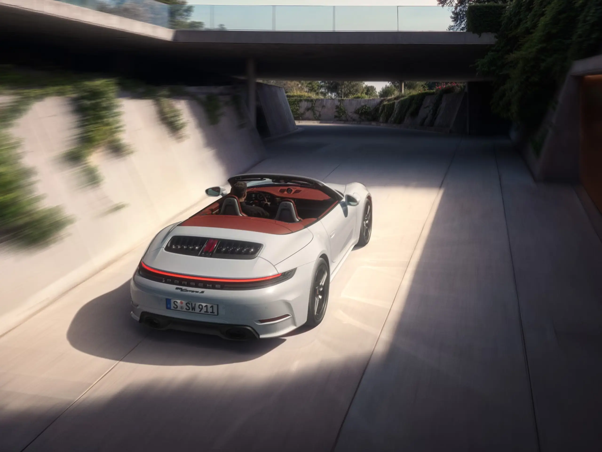 En hvid Porsche 911 Carrera S cabriolet med taget nede kører gennem en moderne, solbelyst tunnel omgivet af betonvægge med grøn beplantning.