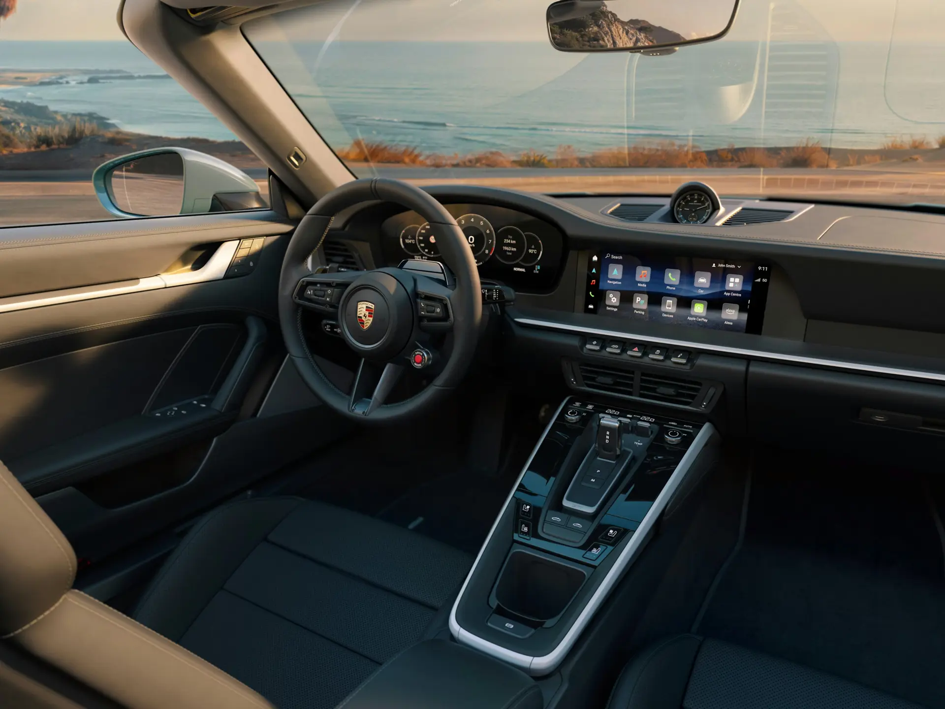 Interiør i en moderne Porsche 911 Carrera 4S med fokus på cockpit, rat og digitalt infotainmentsystem ved kystvejen.