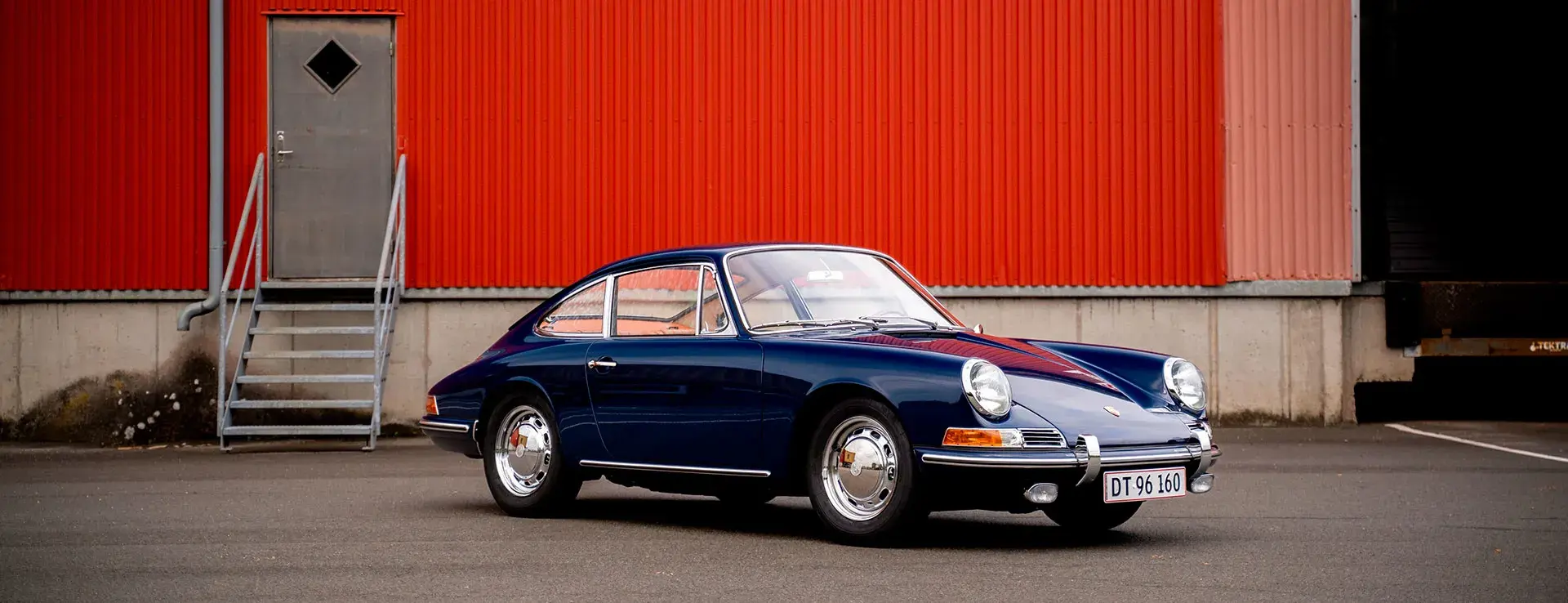 Mørkeblå klassisk Porsche 911 parkeret foran en rød industribygning.