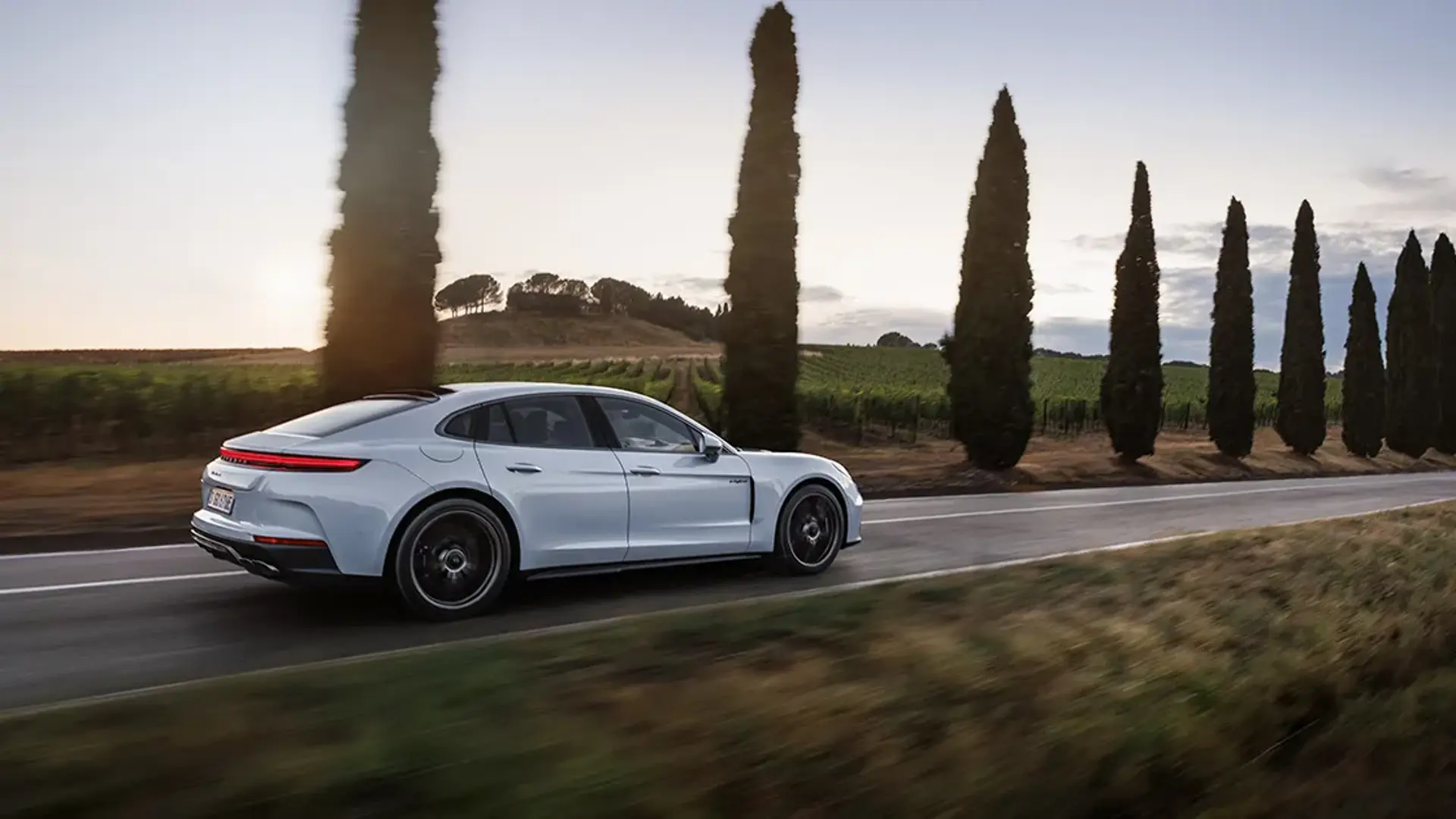 En hvid Porsche Panamera kører ad en landevej omgivet af vinmarker og slanke cypresser i aftenskumringen.