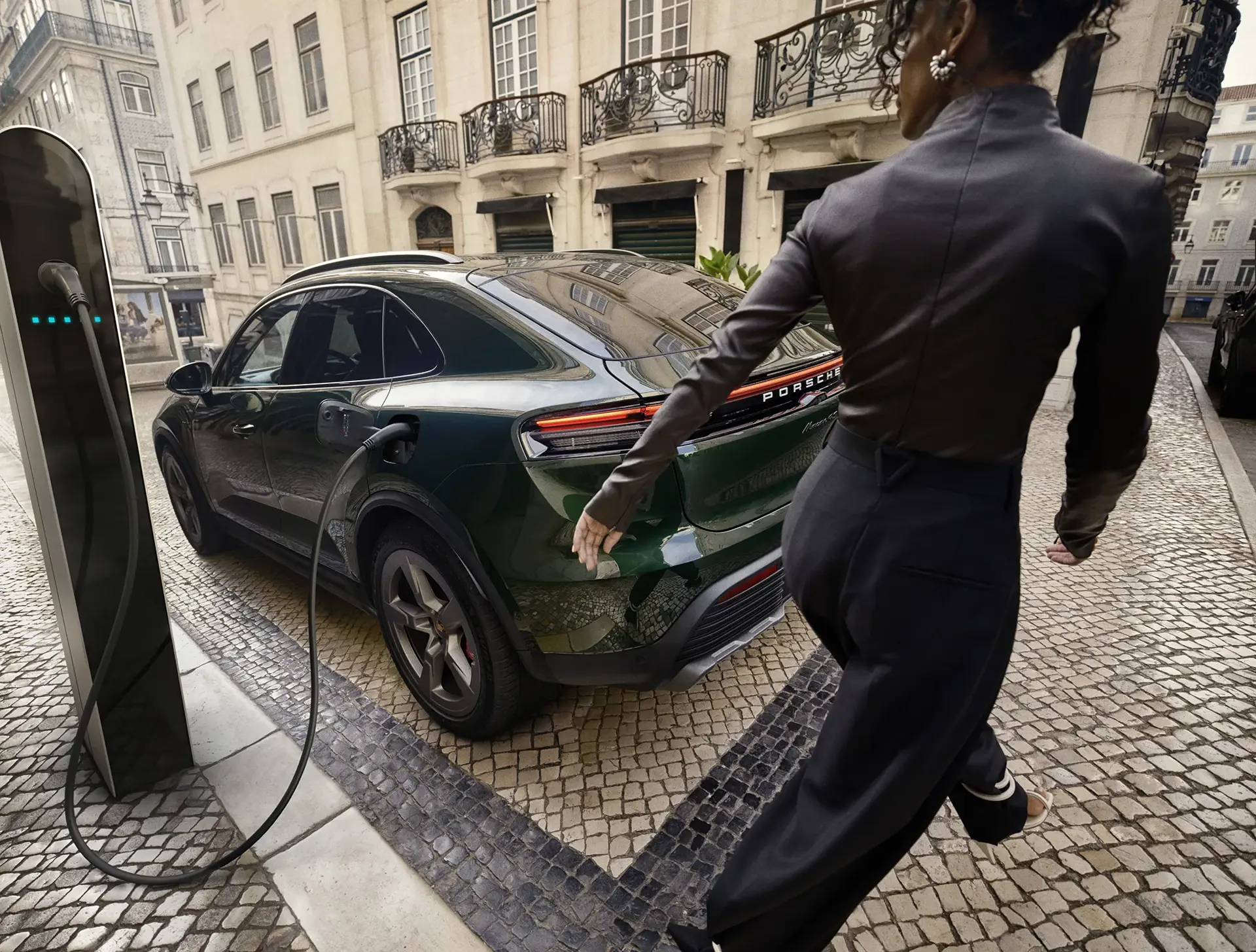 En kvinde går forbi en mørkegrøn elbil fra Porsche, der står til opladning ved en ladestander i en brostensbelagt bygade.