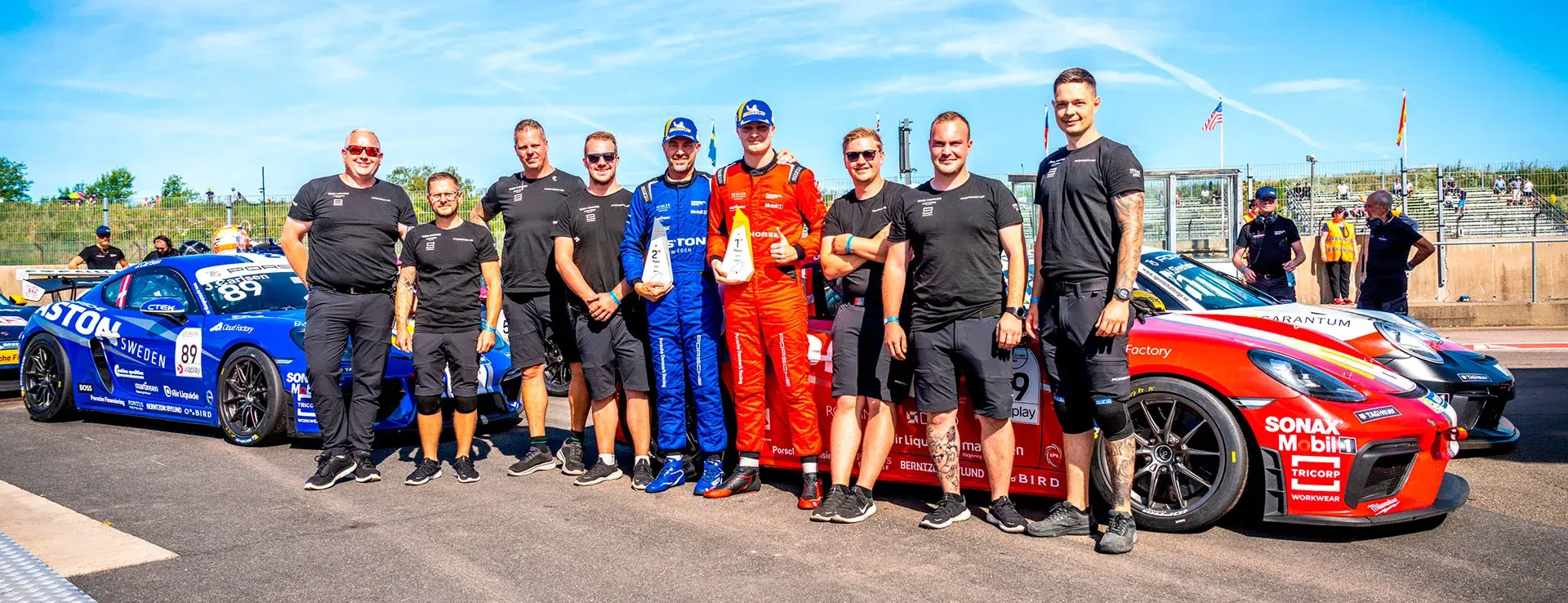 Racerførere og teammedlemmer står foran deres Porsche-løbsbiler på pitlane ved Falkenbergbanen efter et løb.