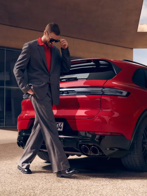 En mand i jakkesæt går forbi bagenden af en rød Porsche Cayenne GTS parkeret ved en moderne bygning.