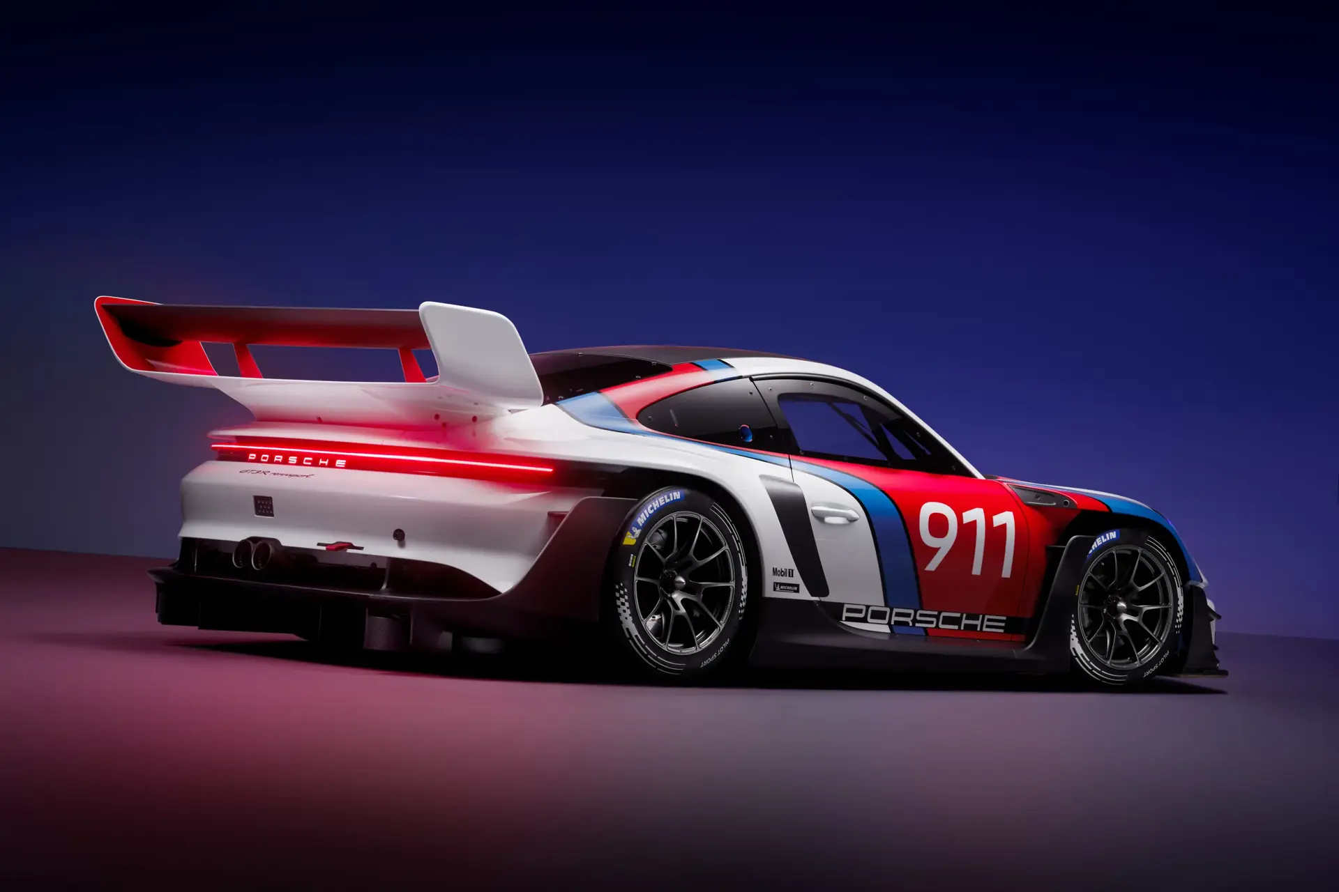En hvid, rød og blå Porsche 911 GT3-racerbil set skråt bagfra i et studiemiljø med mørk baggrund.