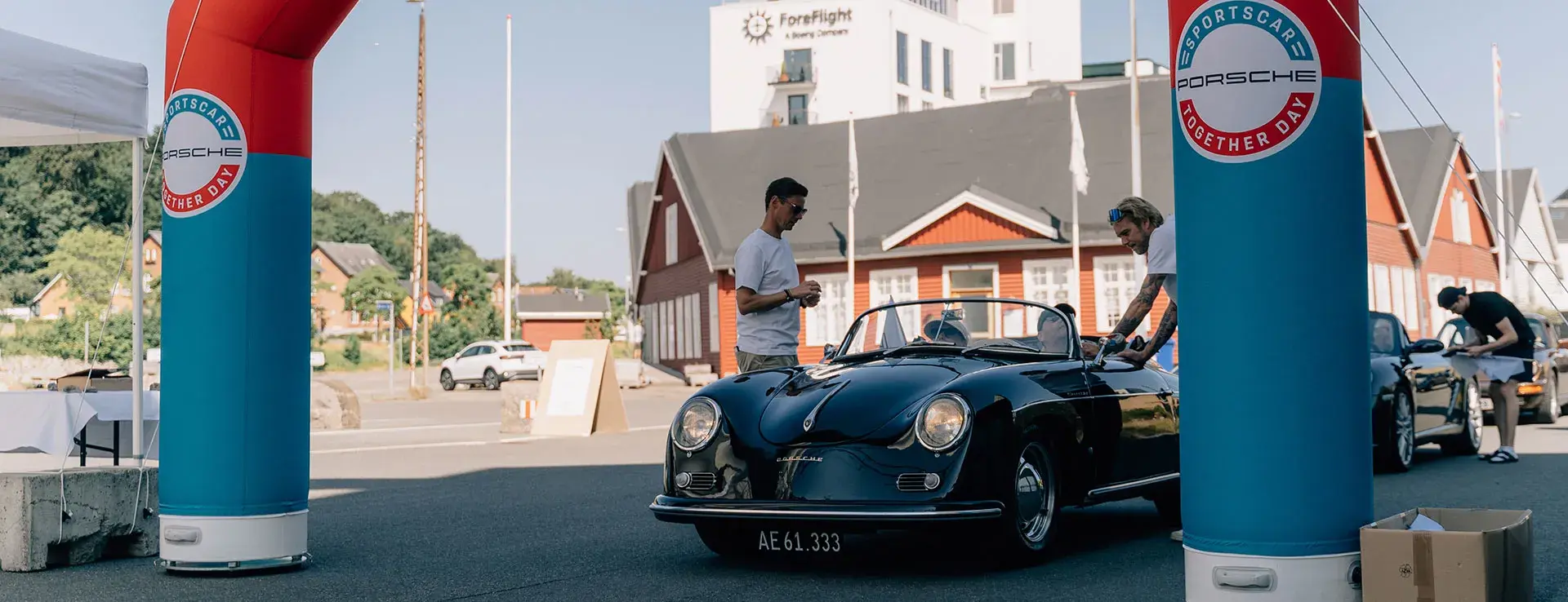En sort klassisk Porsche cabriolet ankommer til et Porsche Sports Car Together Day-event mellem to blå og røde oppustelige portaler foran røde træhuse.