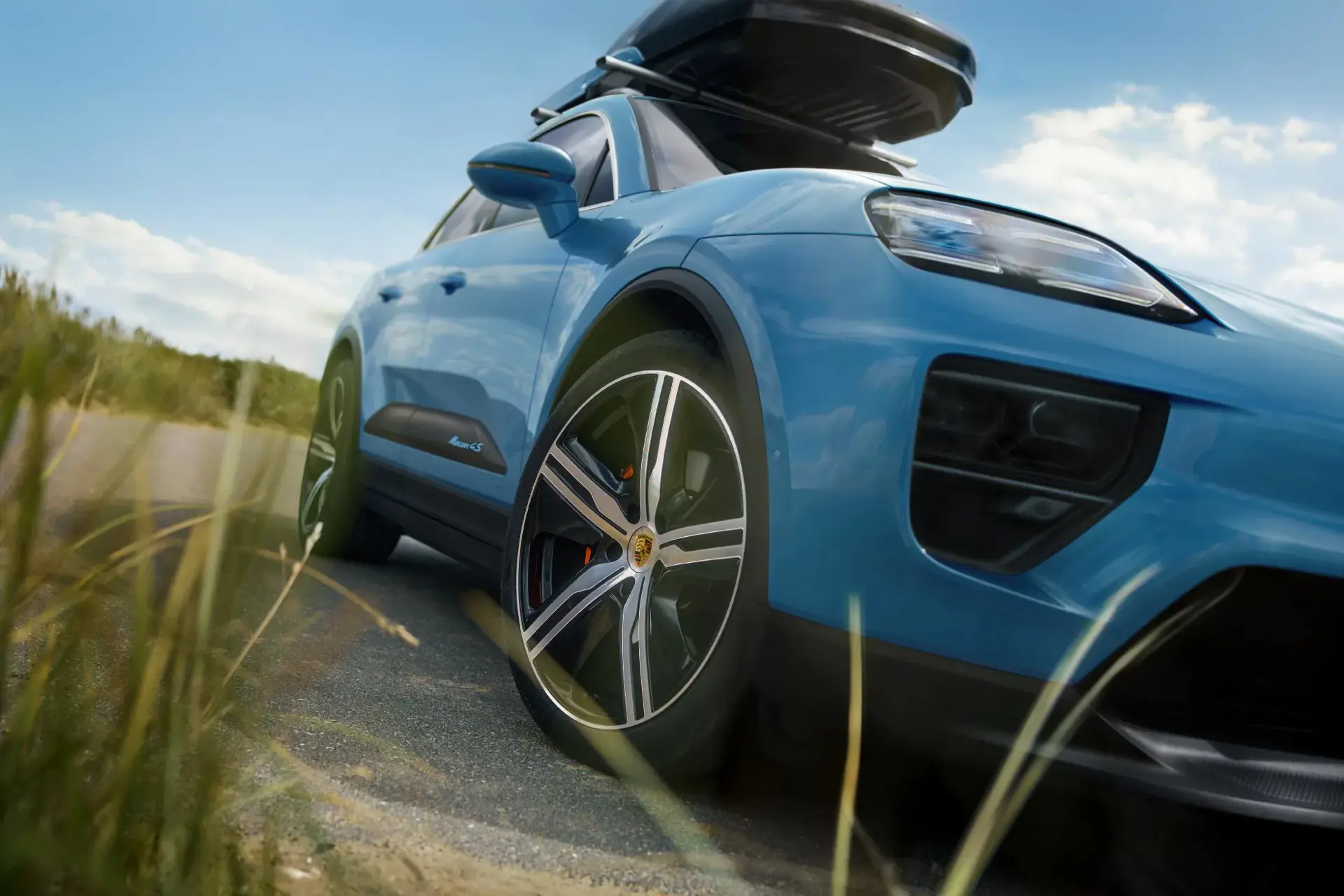 Blå Porsche Macan set nedefra fra siden med tagboks monteret, parkeret på en landevej under blå himmel.