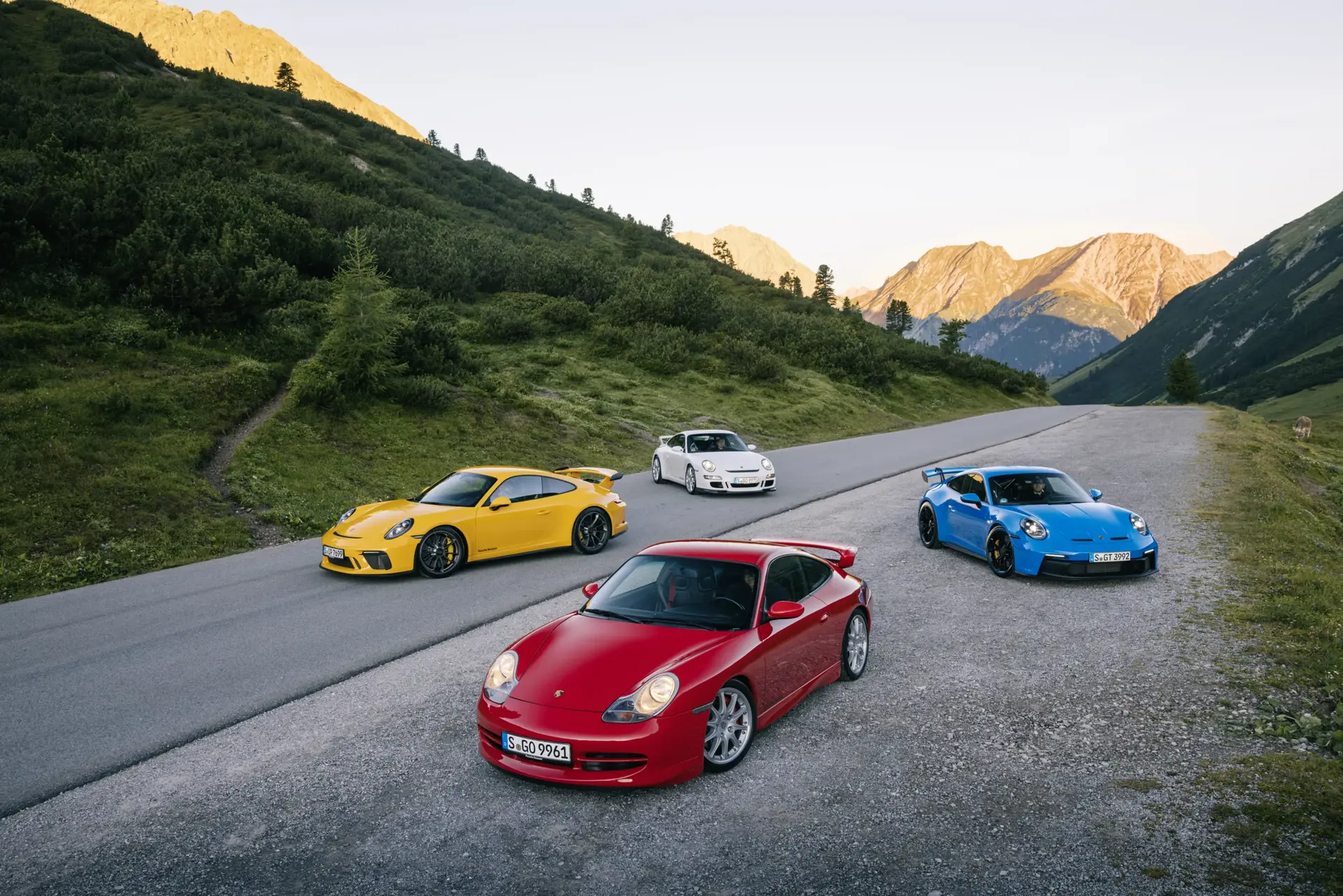 Fire farverige Porsche 911 GT3-biler holder parkeret på en bjergvej i et grønt alpelandskab ved solnedgang.