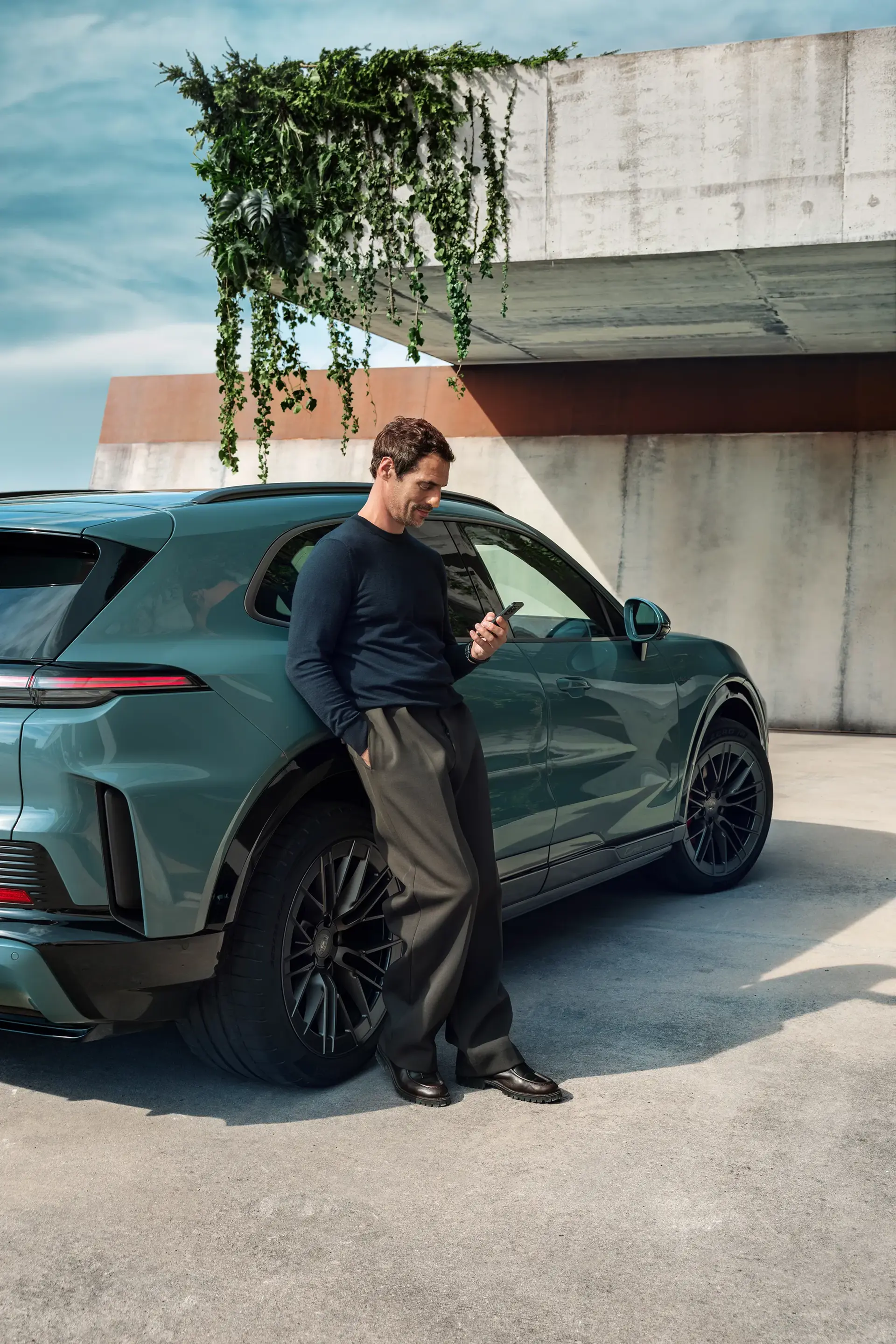 En mand står og kigger på sin smartphone, mens han læner sig op ad en grøn elektrisk Porsche Cayenne under en moderne betonbygning med hængende planter.
