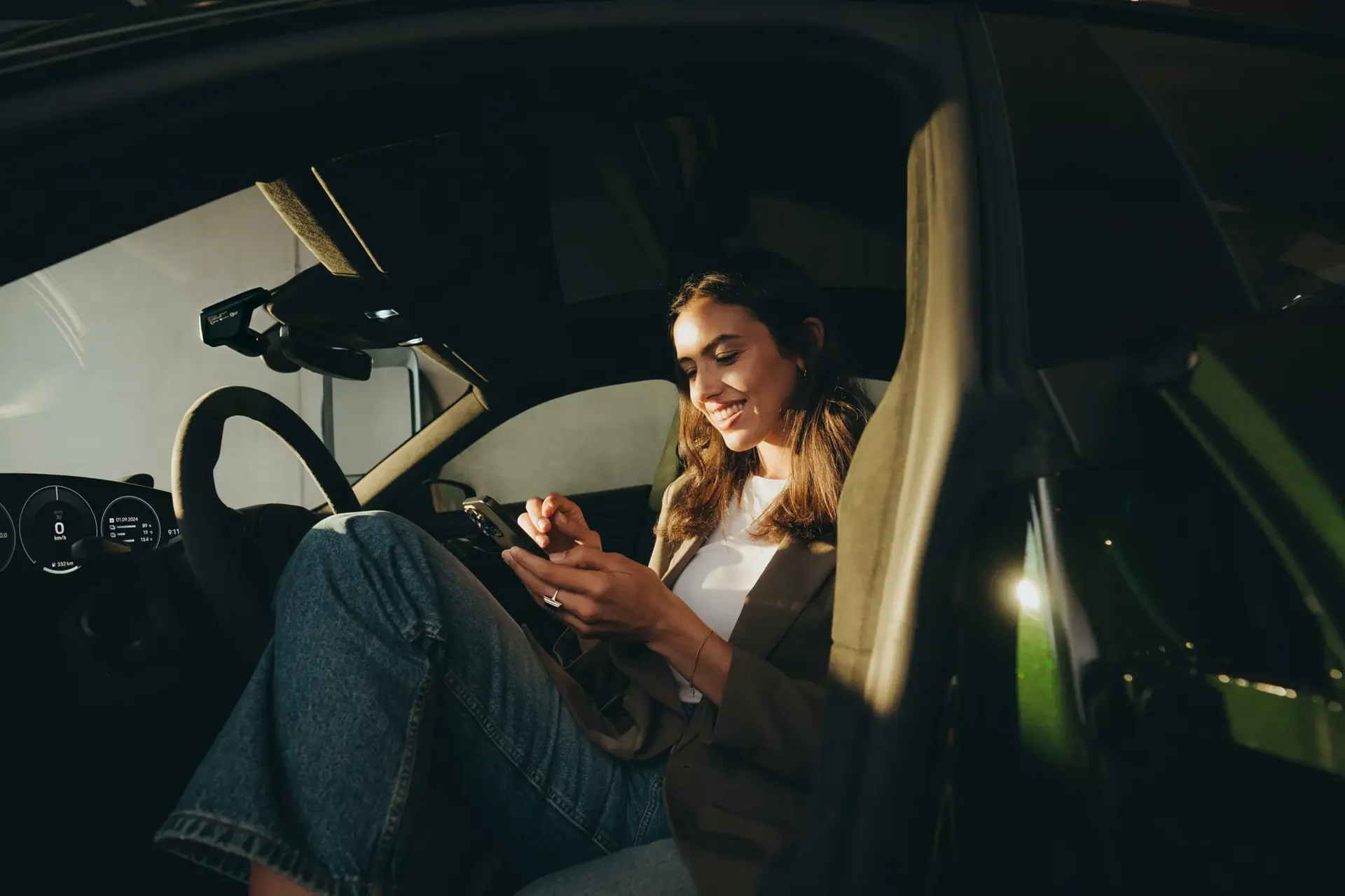 Ung kvinde sidder i en Porsche med åben dør og bruger en app på sin smartphone.