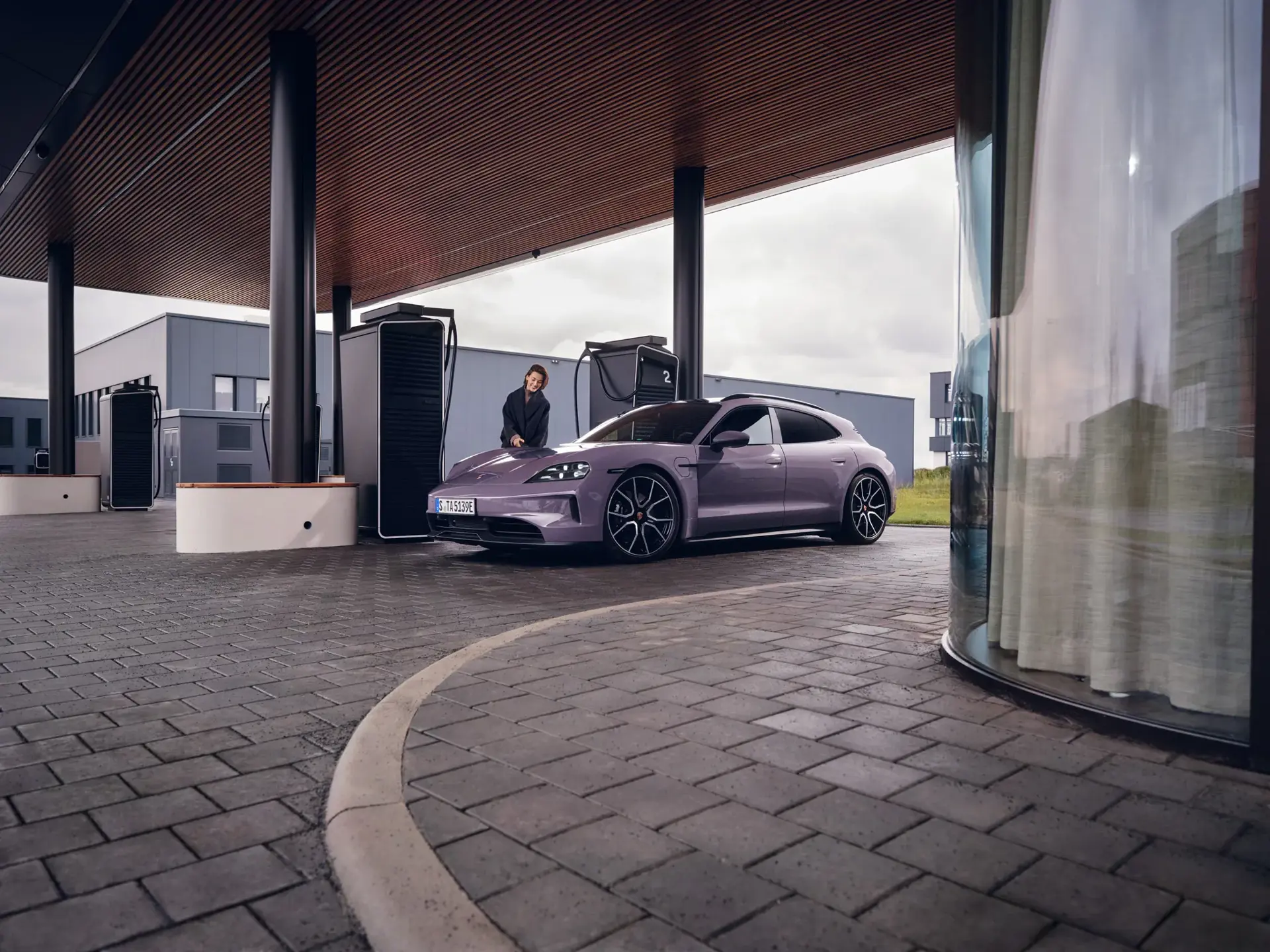 En lyserød Porsche Taycan holdes til opladning ved en moderne el-ladestation, mens en person står lænet op ad bilen.