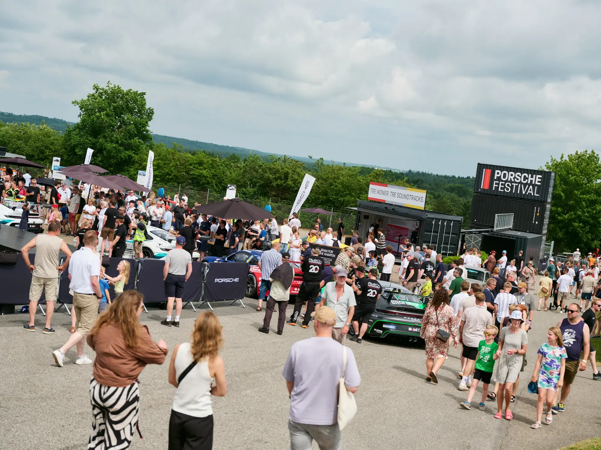 Mange mennesker går rundt og ser på udstillede Porsche-biler ved et udendørs Porsche-festivalarrangement.