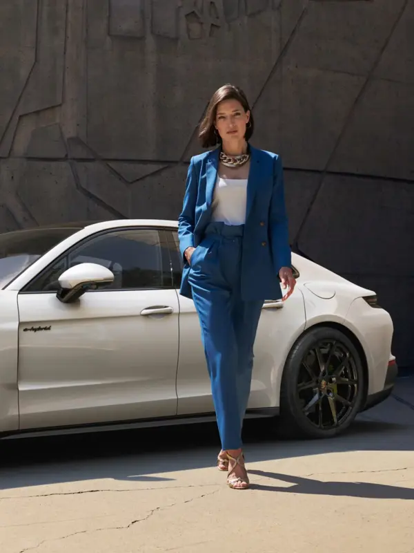 En person i blå habit går foran en hvid Porsche Panamera E-Hybrid parkeret ved en rå betonvæg.
