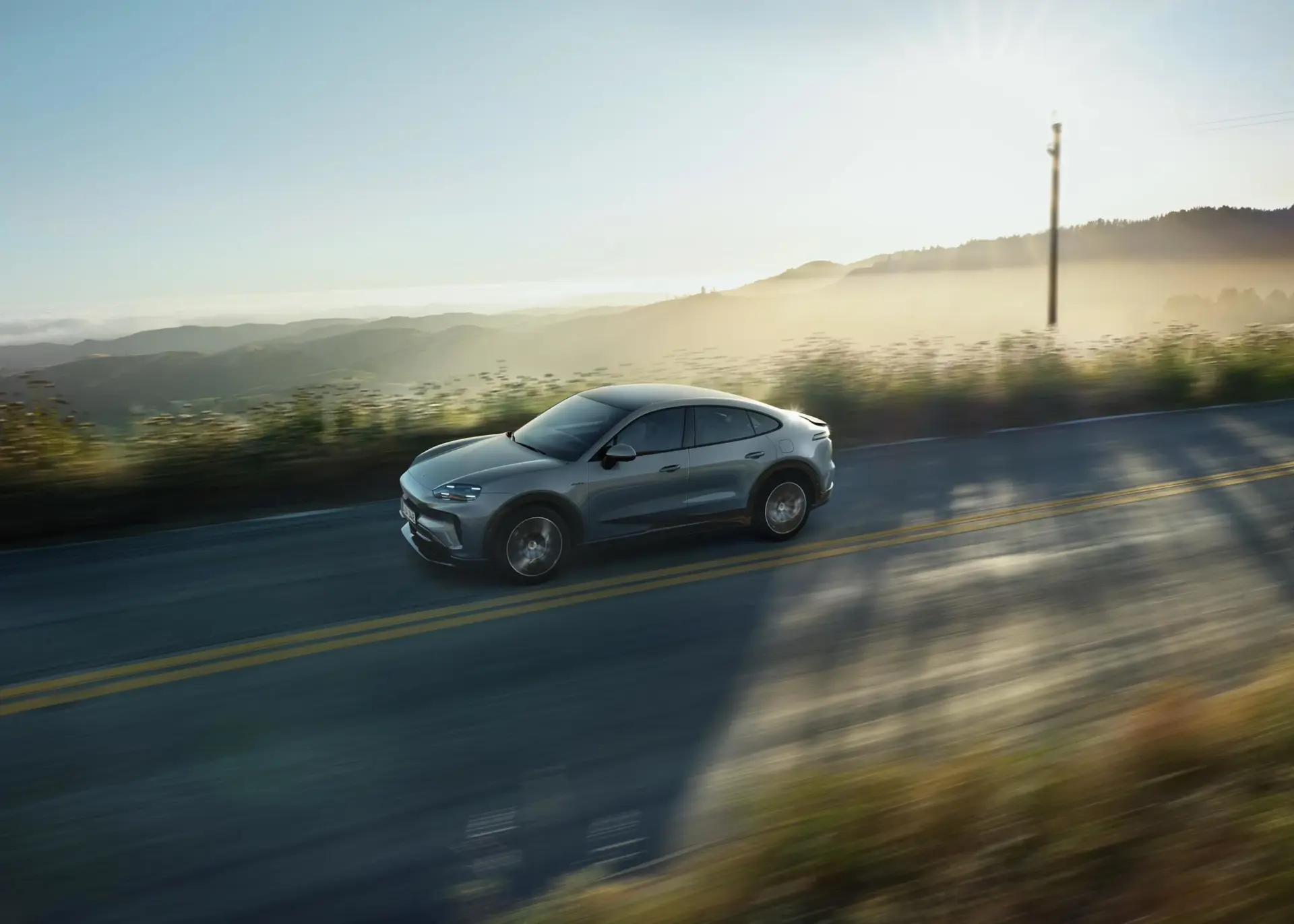 En grå, elektrisk Cayenne coupé kører på en landevej gennem et kuperet landskab i morgensol.