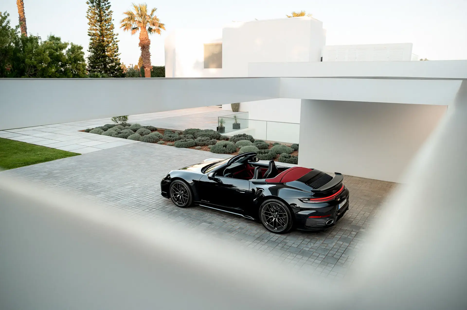 Sort Porsche 911 Cabriolet parkeret i en moderne indkørsel foran en hvid minimalistisk villa med palmer i baggrunden.