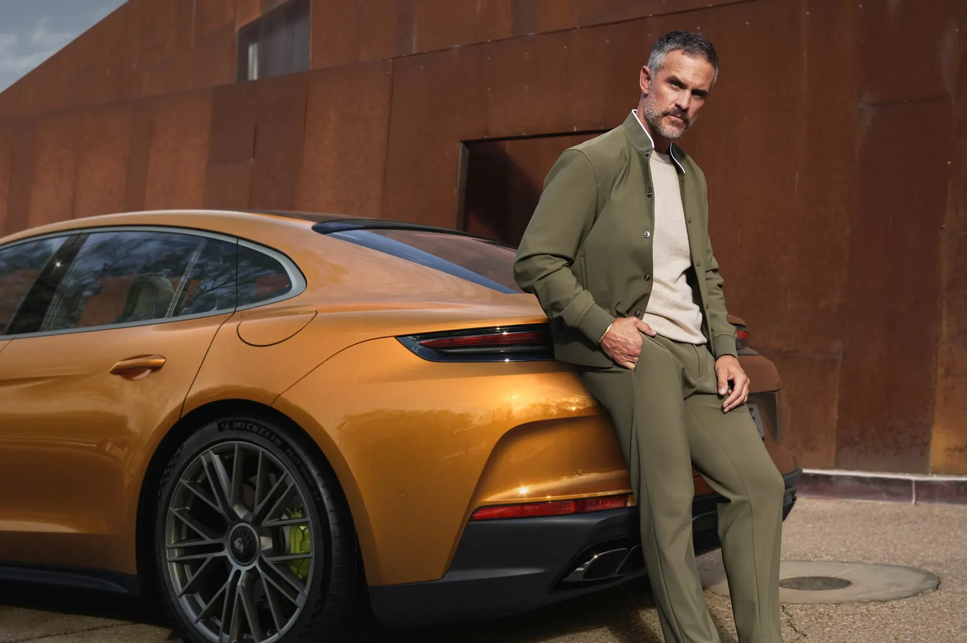Mand i grøn dragt læner sig op ad en gylden Porsche Panamera foran en rustfarvet væg.
