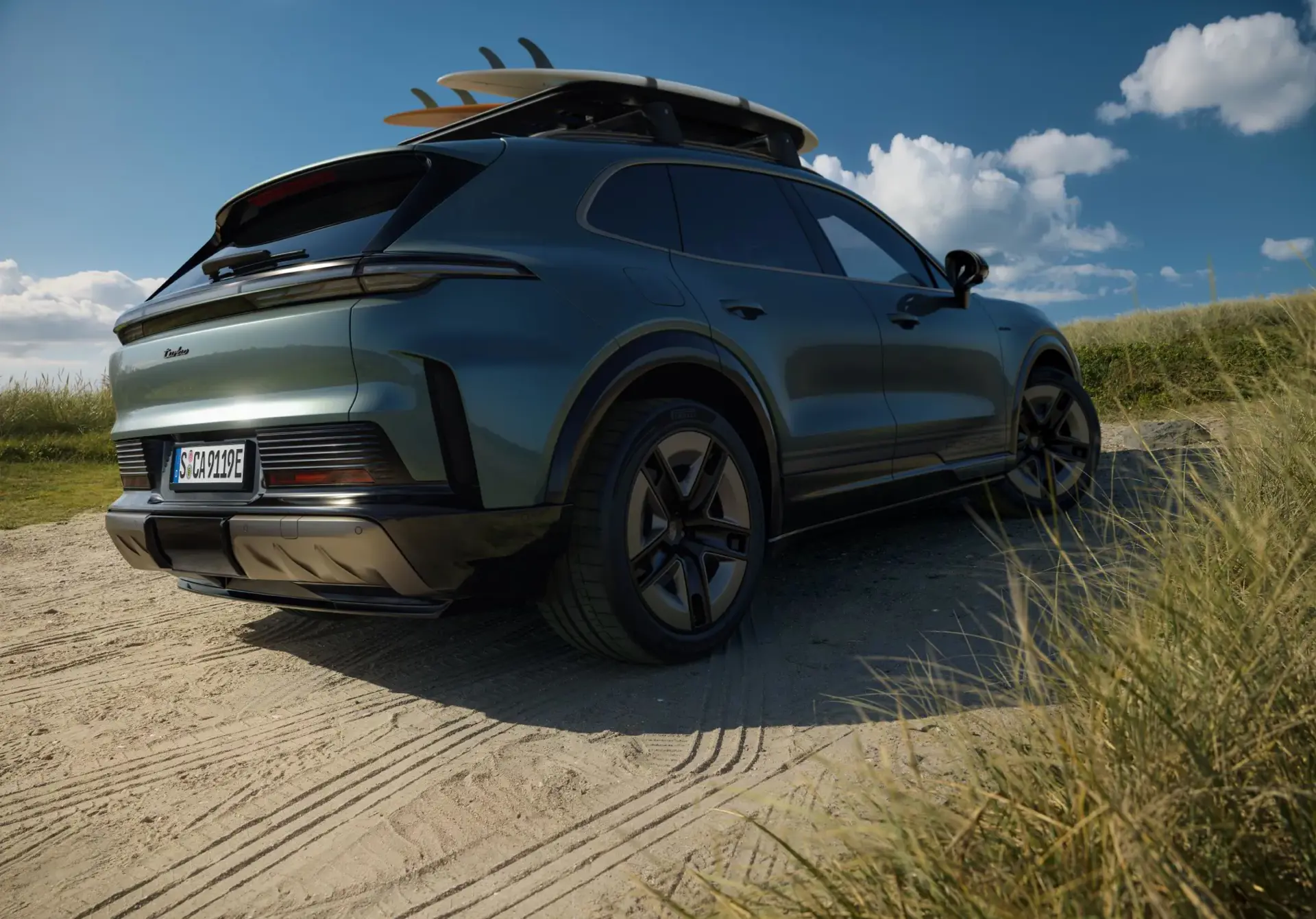 En mørkegrøn elektrisk Porsche Cayenne med surfbrætter på taget holder parkeret på en sandvej i klitterne under en blå himmel.