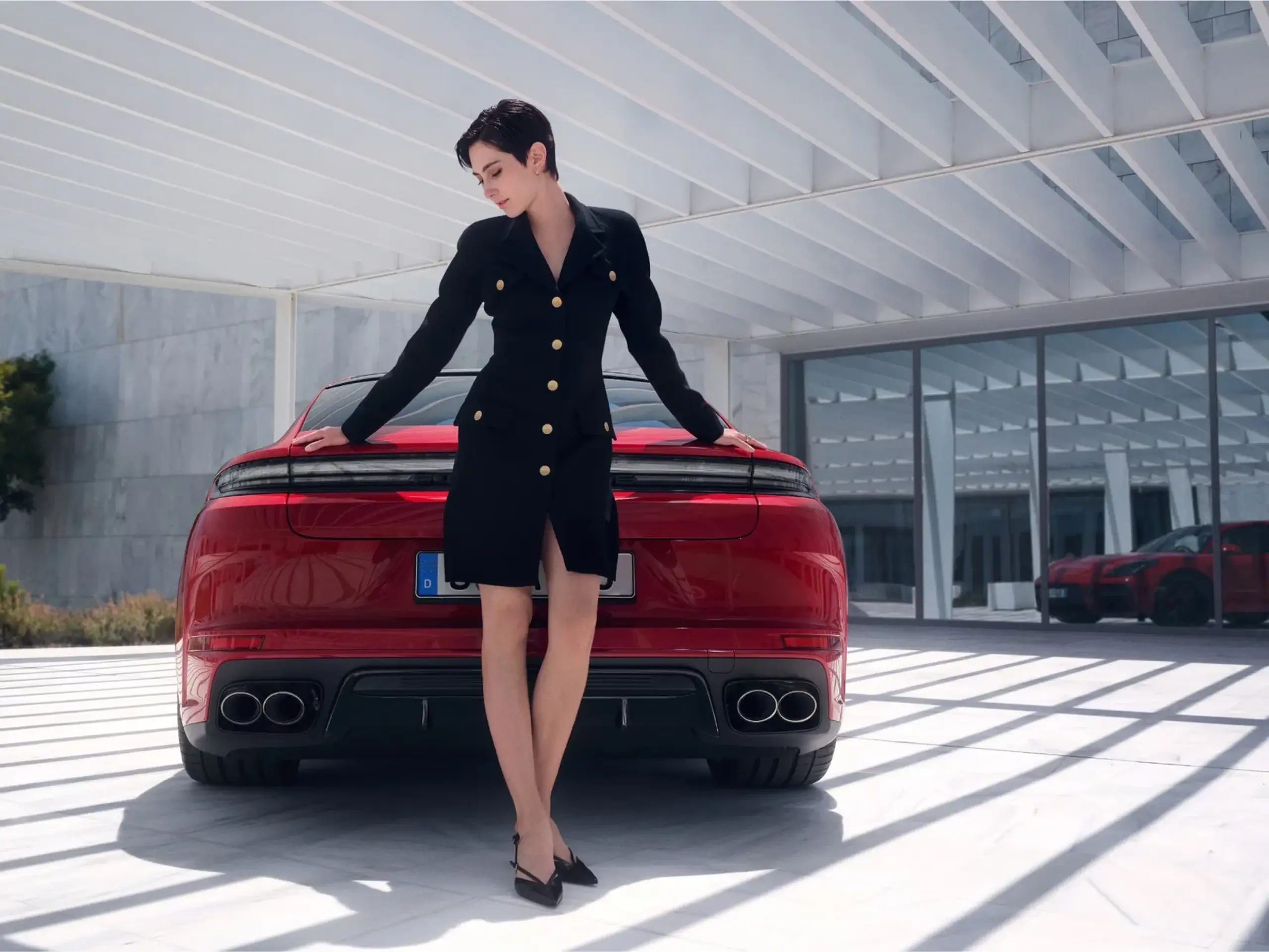 En kvinde i sort blazer-kjole står lænet op ad bagenden af en rød Porsche Panamera GTS i en moderne, lys arkitektonisk ramme.