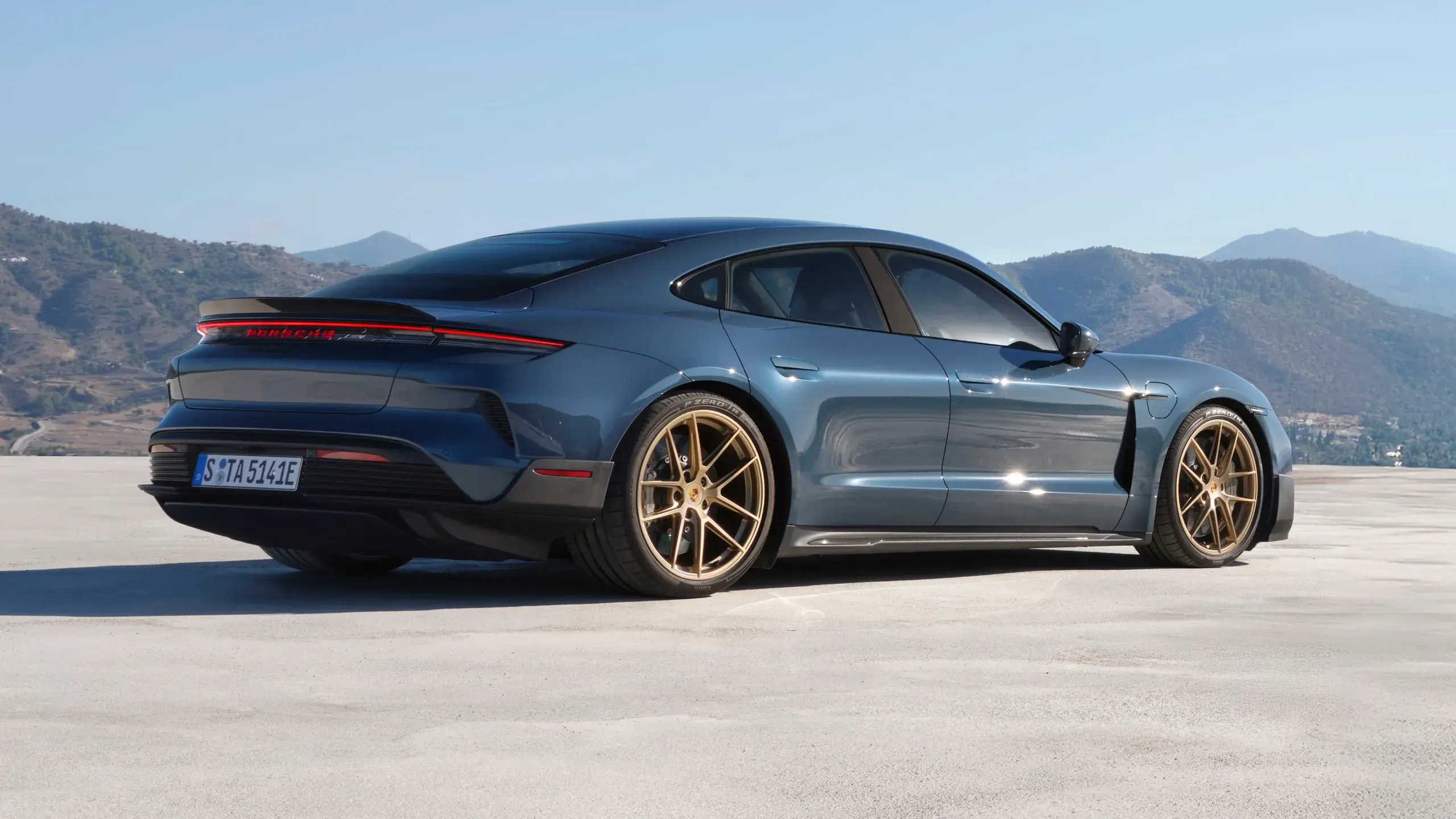 Mørkeblå sportsvogn, formentlig en Porsche Taycan, ses i fart på en åben vej under blå himmel.