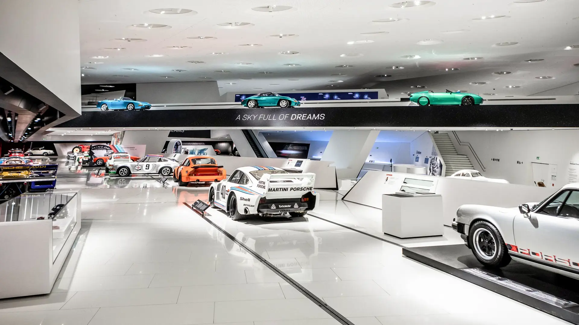 Udstillingshal i et bilmuseum med klassiske og moderne sportsvogne, herunder flere Porsche-modeller, opstillet på rækker under skiltet “A Sky Full of Dreams”.