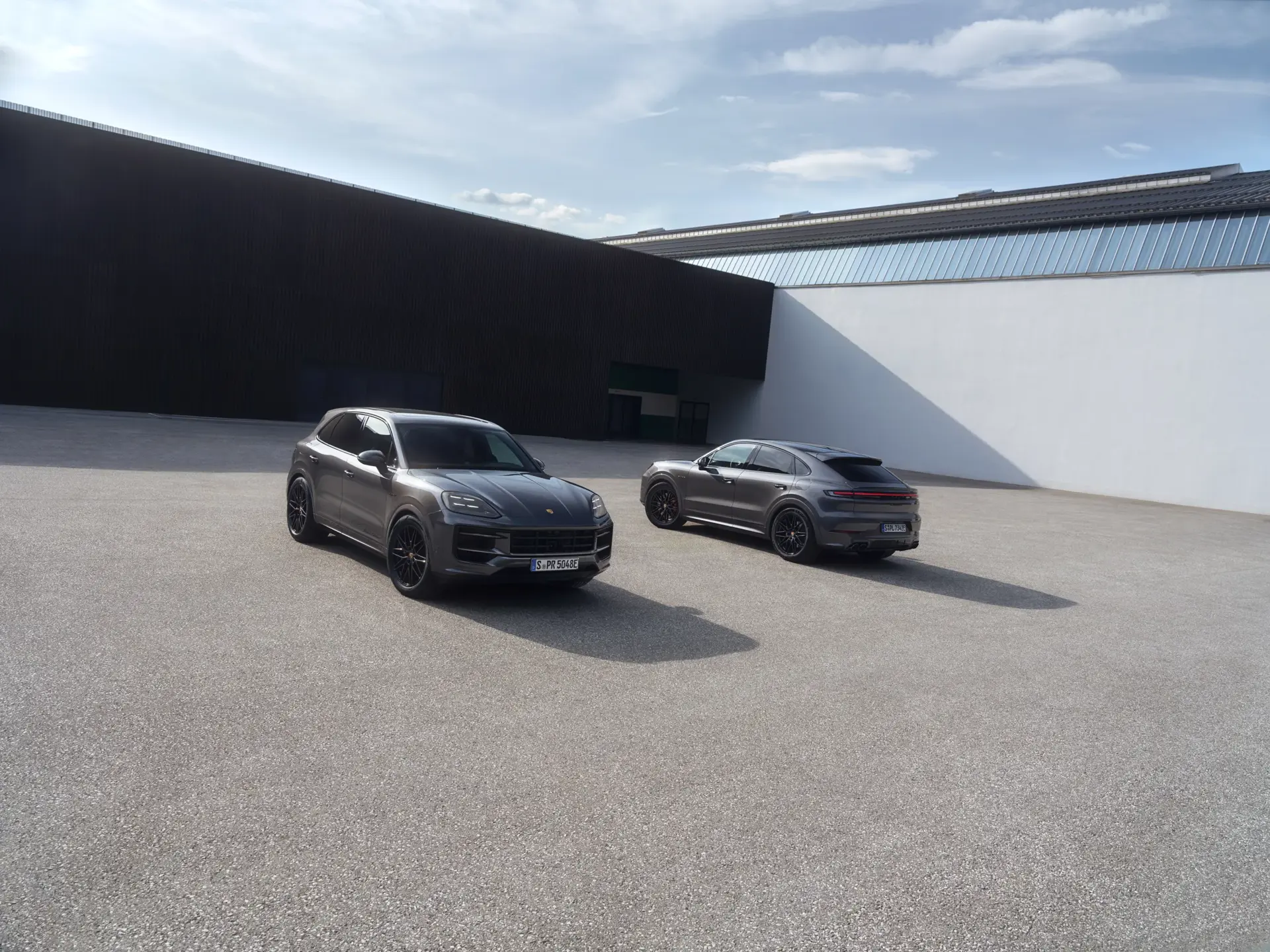 To grå Porsche Cayenne Black Edition SUV’er holder parkeret på en åben, moderne plads foran en minimalistisk bygning.