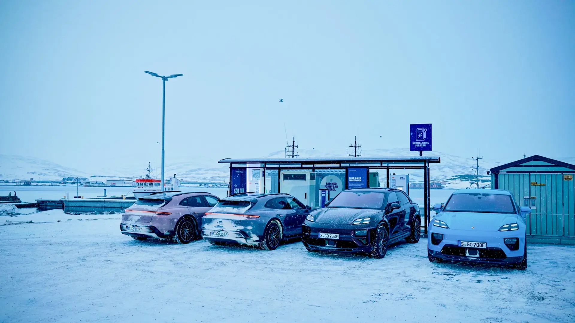 Fire elbiler holder parkeret ved en ladestation i et sneklædt havnemiljø.