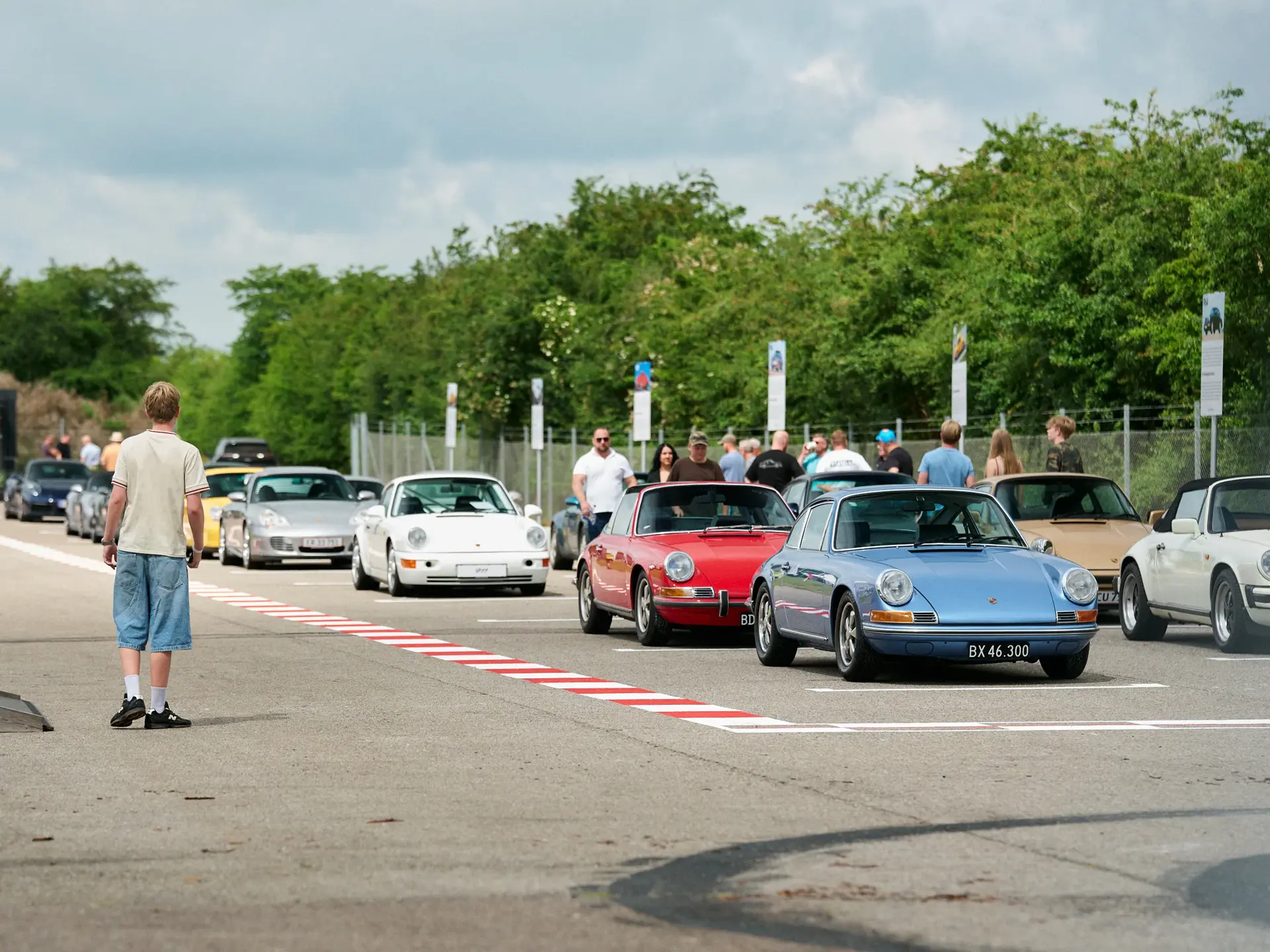 En række klassiske Porsche-sportsvogne holder i kø på Jyllandsringen omgivet af gående tilskuere.