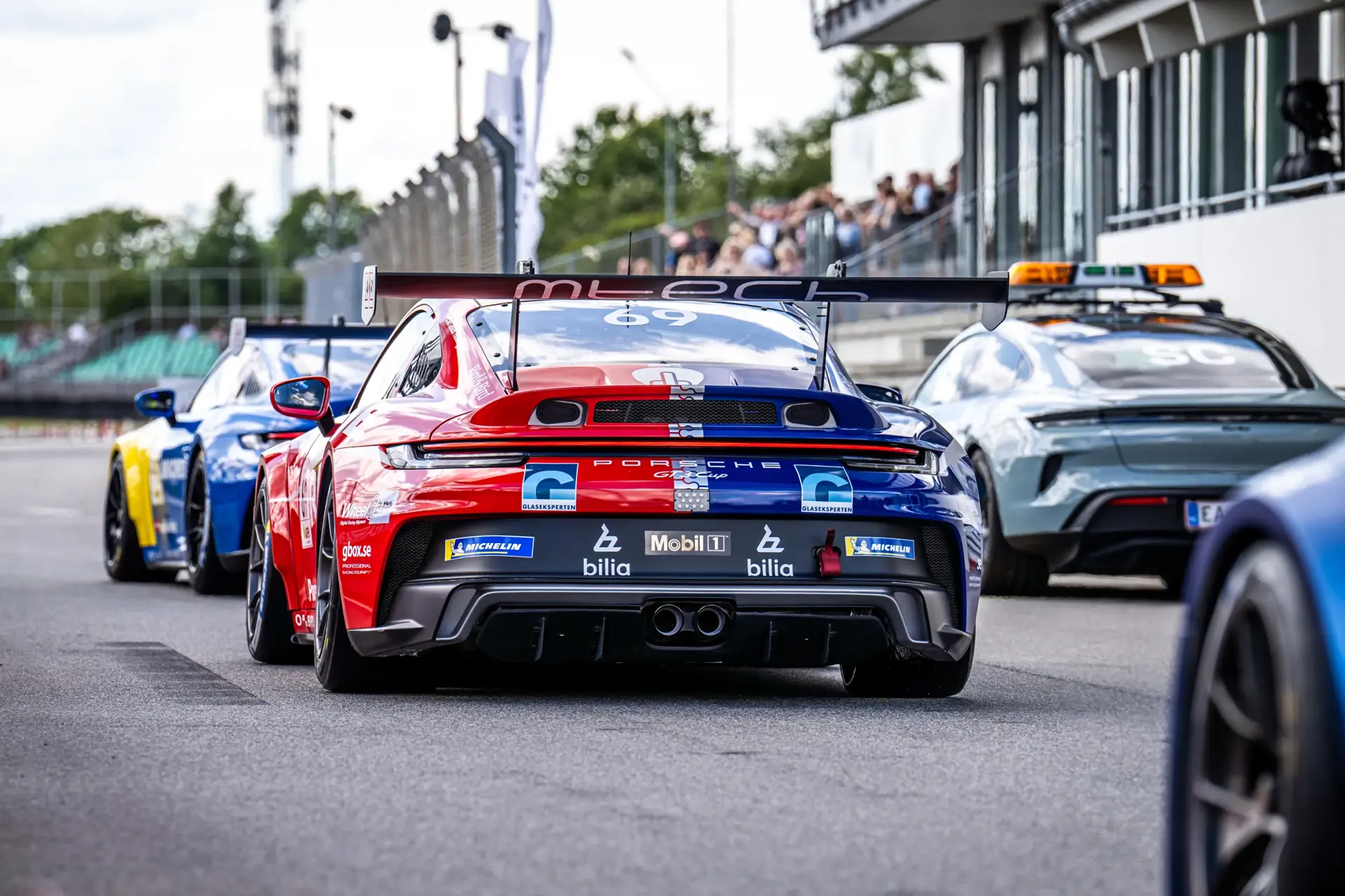 Racerbiler holder på række i pitlane på en racerbane, set bagfra med en rød og blå Porsche i fokus.