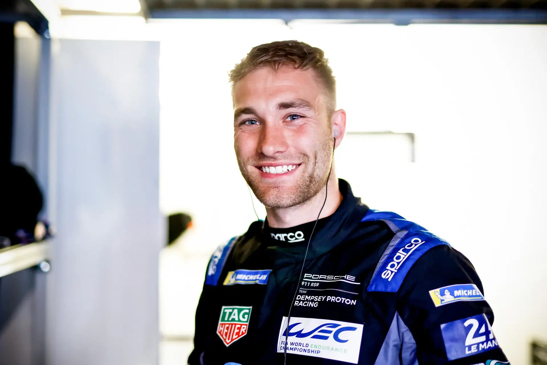 Racerkører i Porsche-holddragt står i en garage og lytter koncentreret med ørepropper.