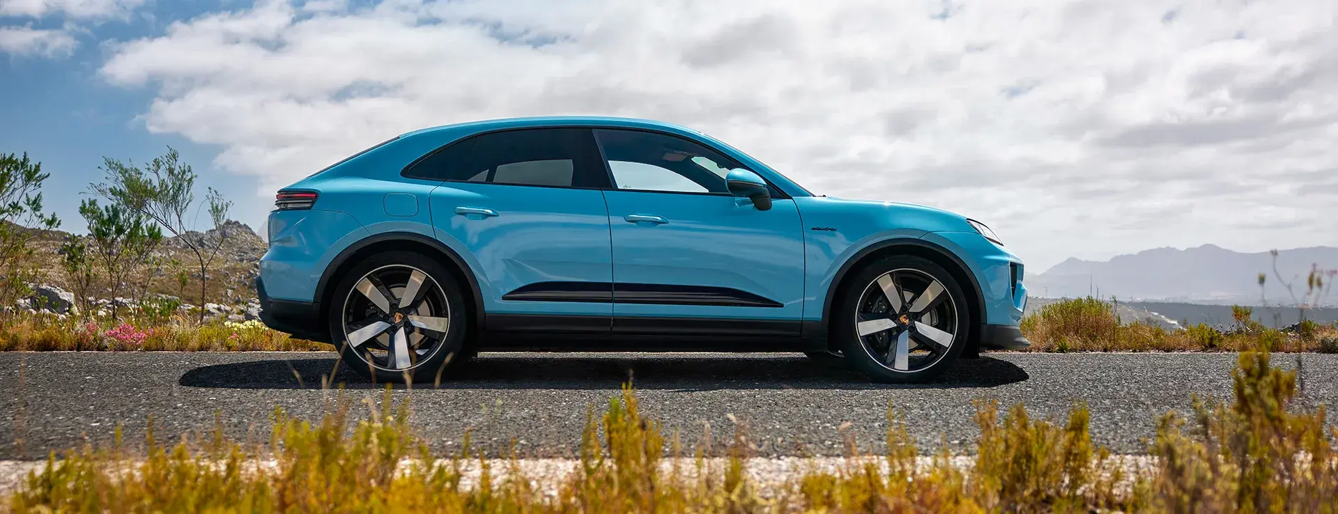 En turkisblå Porsche Macan ses fra siden, parkeret på en landevej i et åbent, bjergrigt landskab under en delvist overskyet himmel.