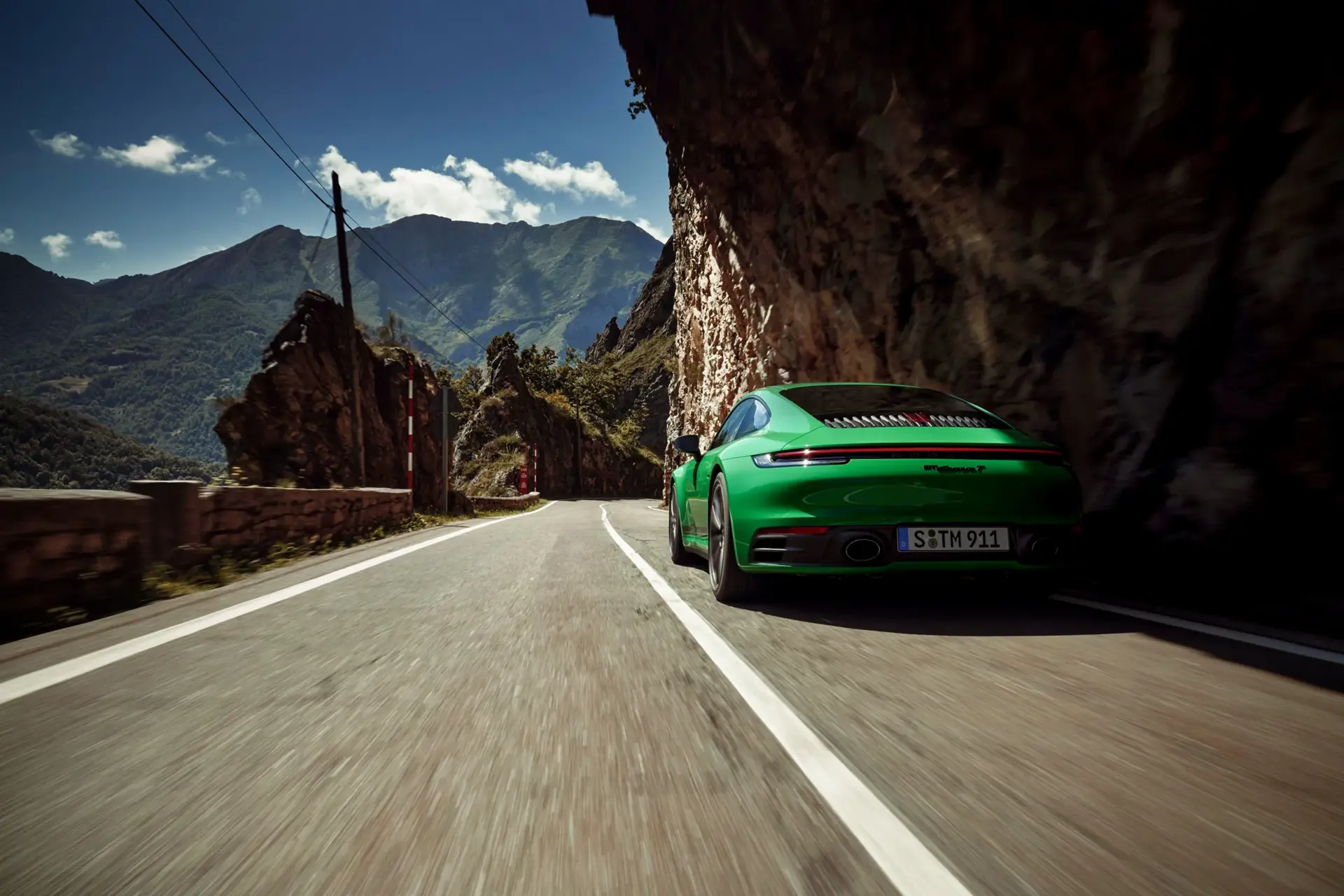 En grøn Porsche 911 Carrera T kører hurtigt gennem en smal bjergvej med klipper og høje bjerge i baggrunden.