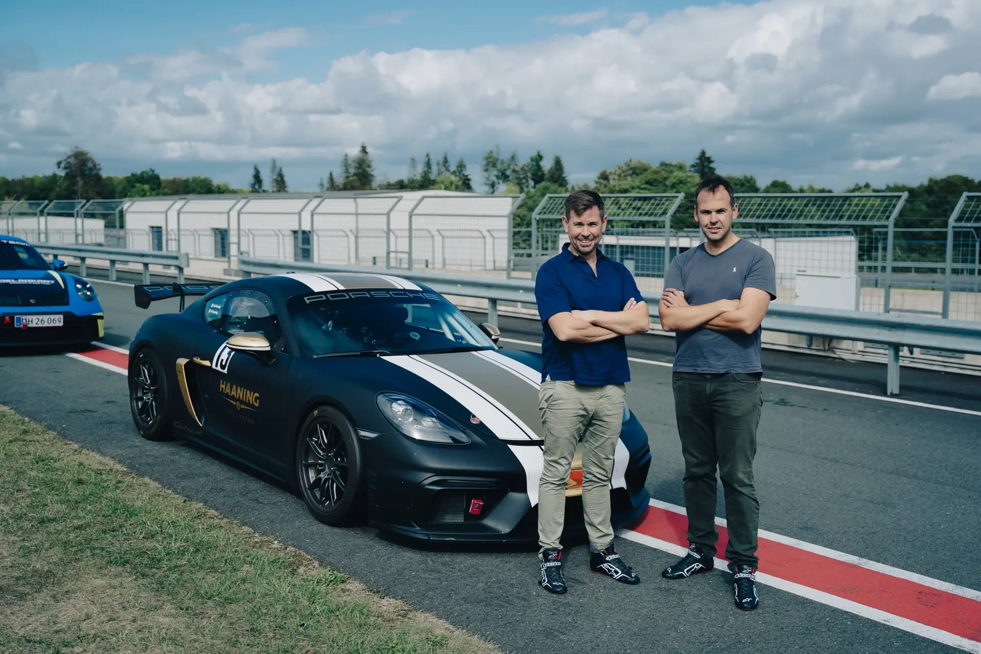 To mænd står foran en sort Porsche racerbil i pitområdet på en racerbane.