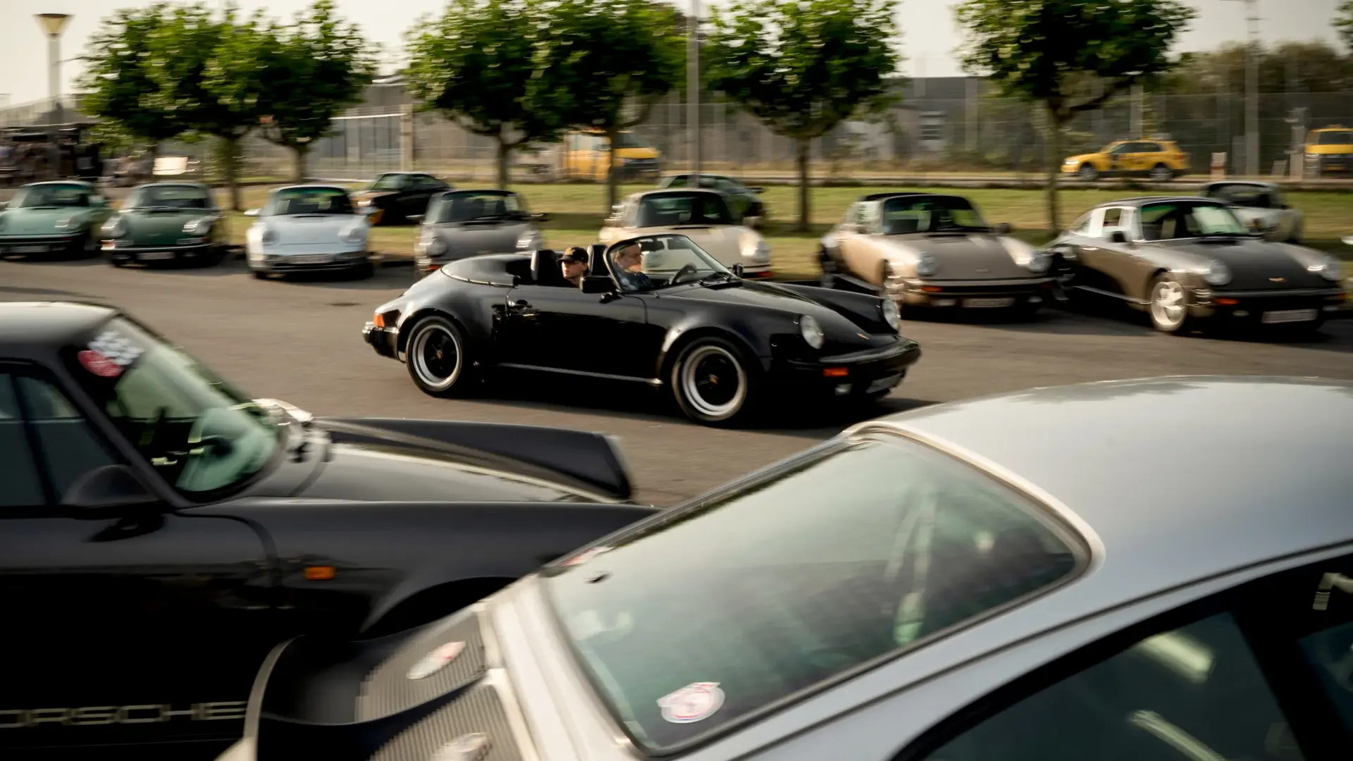 Sort cabriolet sportsvogn kører forbi en parkeringsplads fyldt med klassiske biler i bevægelsesuskarphed.