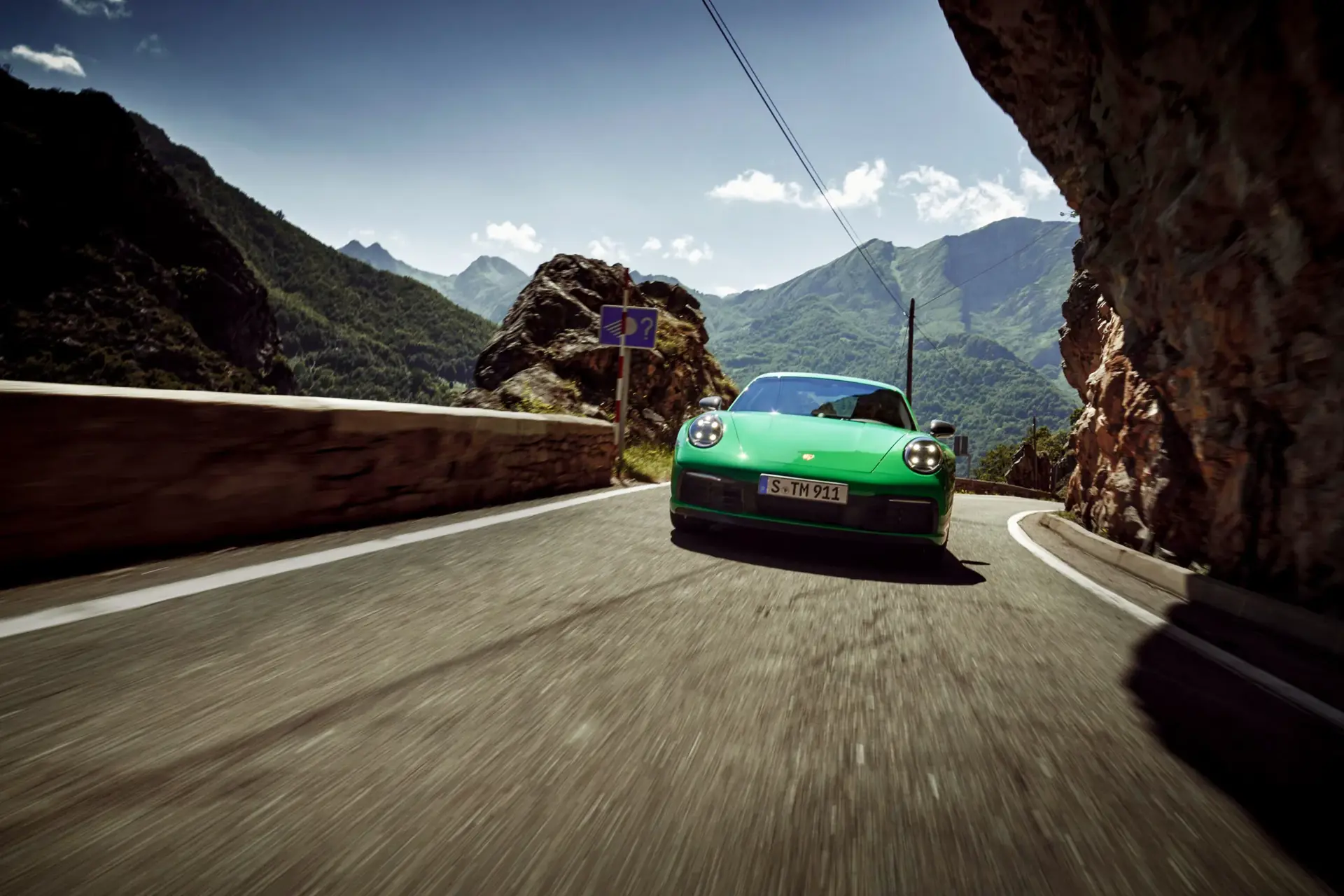 En grøn Porsche 911 Carrera T kører hurtigt gennem en snoet bjergvej omgivet af grønne bjerge.