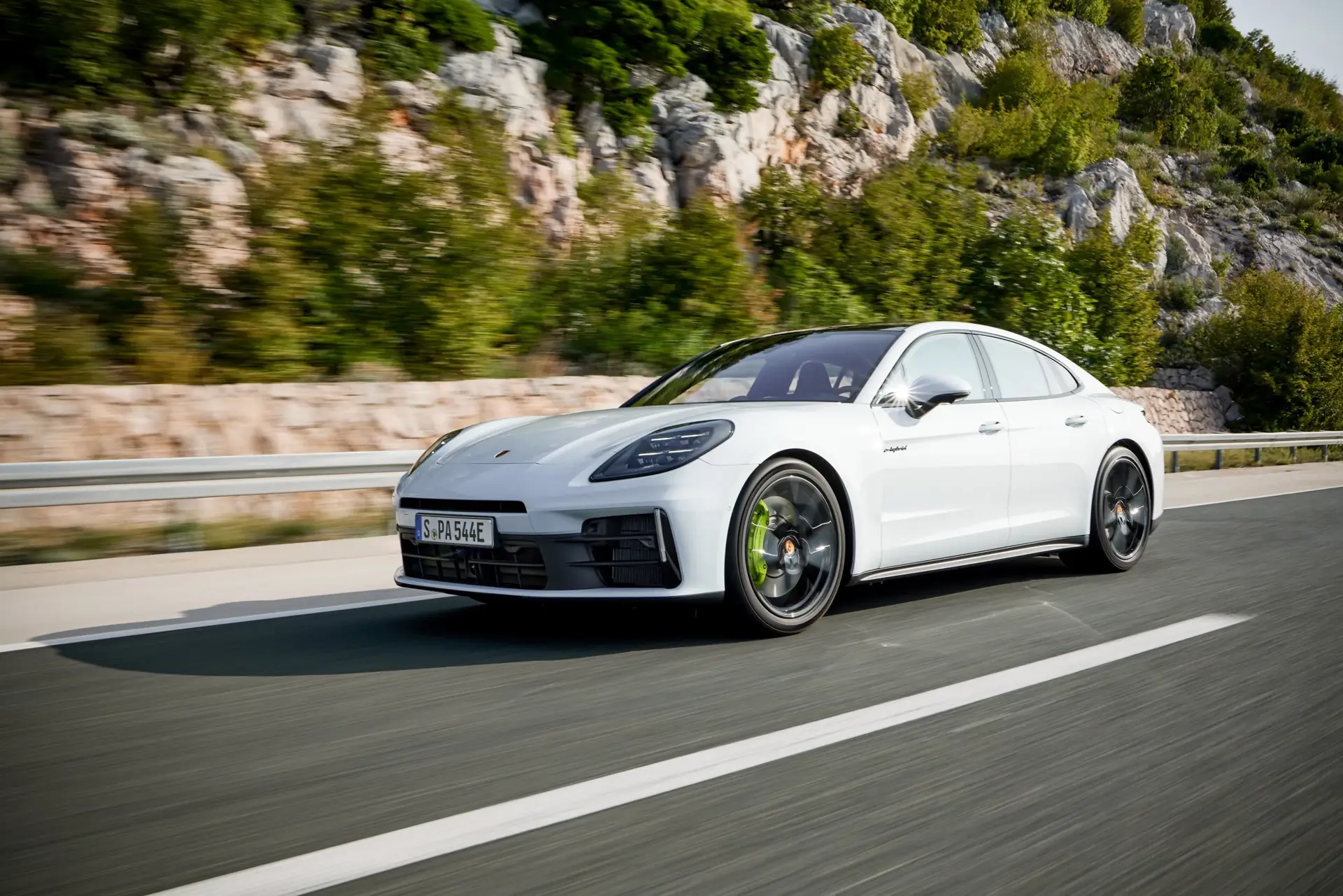 Hvid Porsche Panamera kører på en motorvej med klippefyldt, grønt landskab i baggrunden.