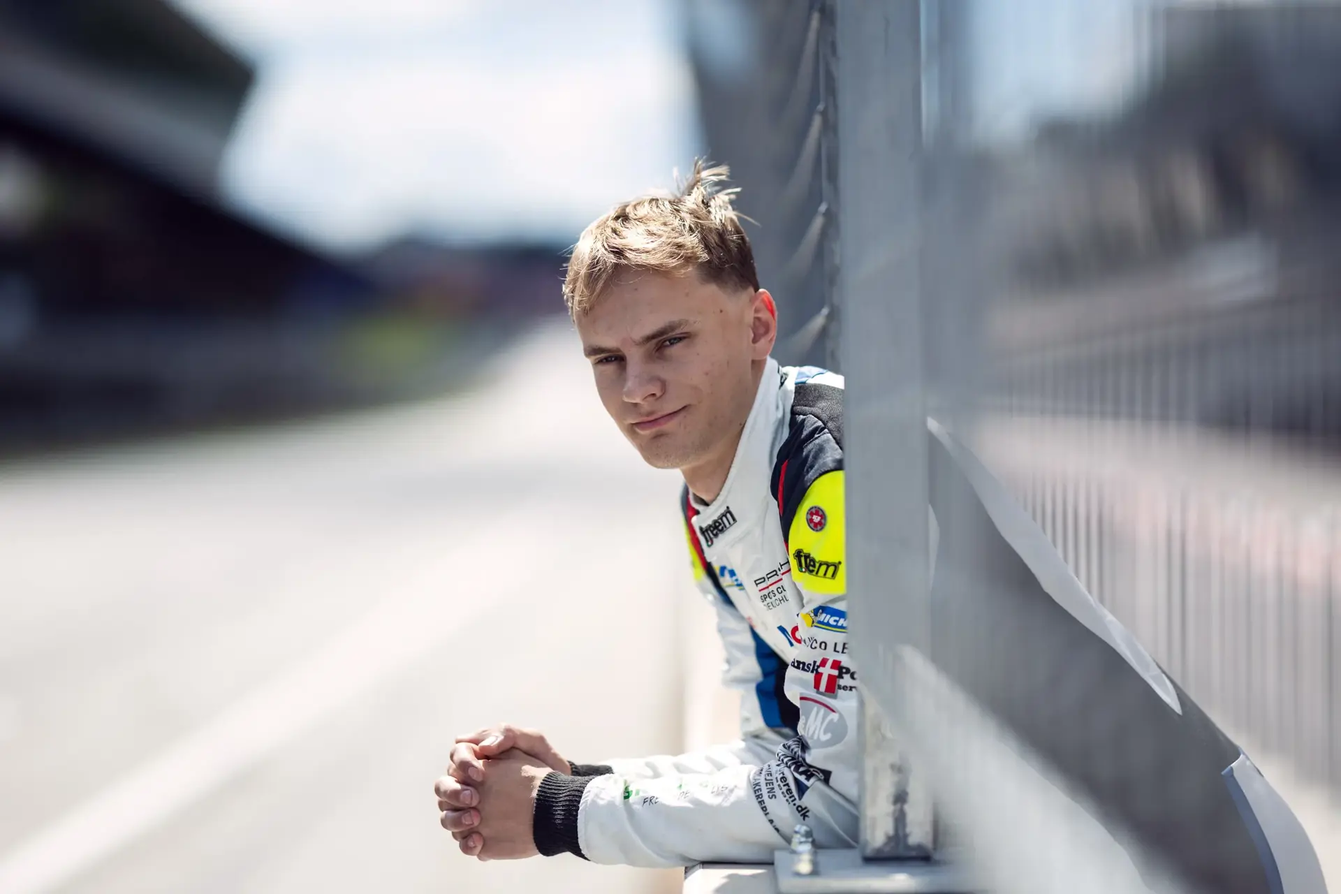 Ung racerkører i køredragt læner sig afslappet mod rækværket ved en tom racerbane.