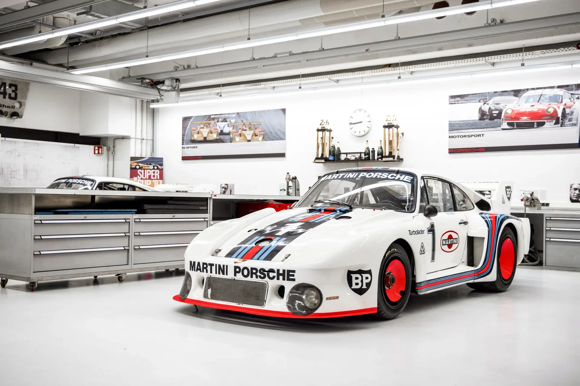 Hvid Porsche-racerbil i Martini-bemaling står i et lyst, moderne værksted omgivet af værkstedsborde og motorsportspokaler.
