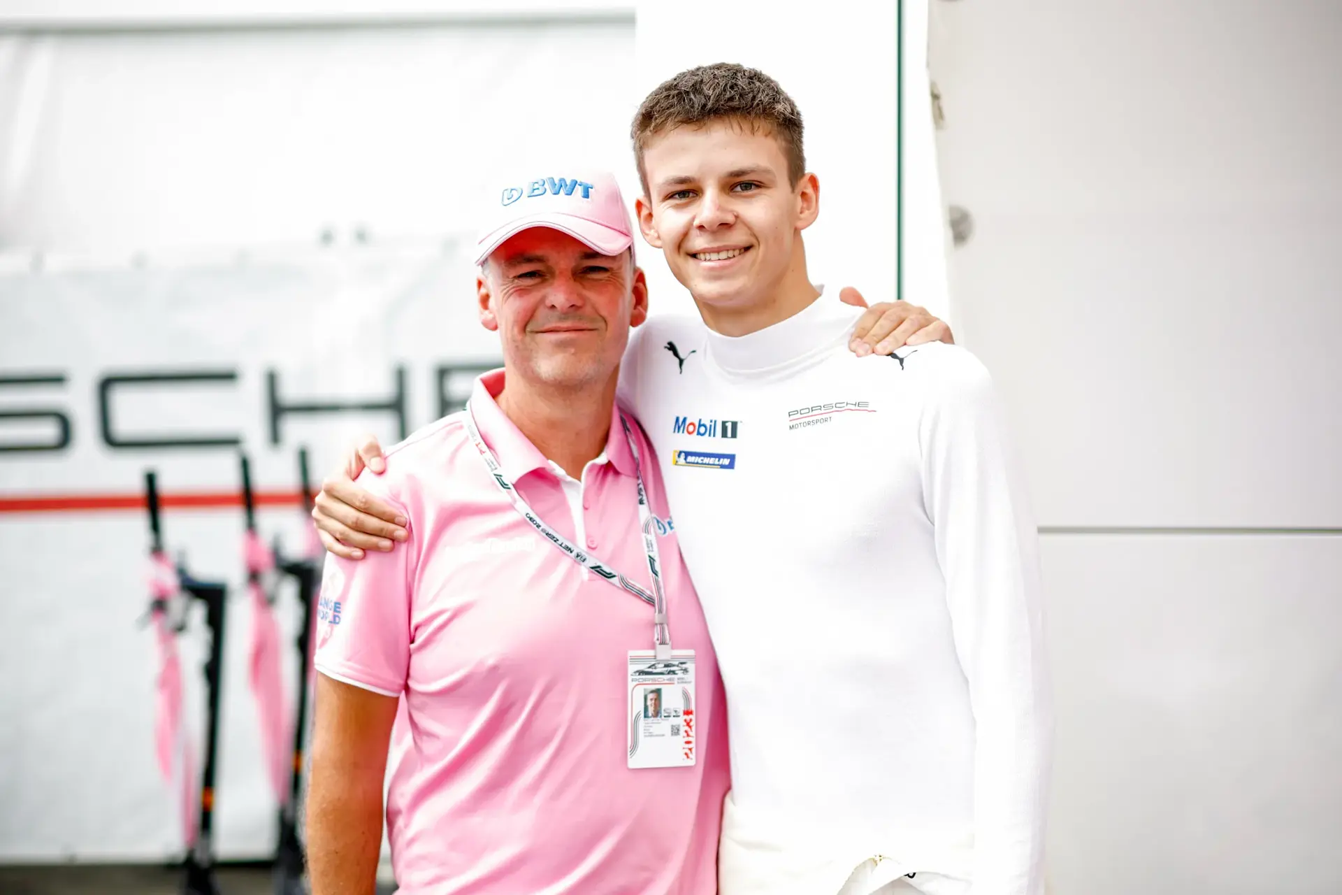 To personer poserer arm i arm foran et Porsche Motorsport-område, hvor den ene bærer lyserød team-beklædning og den anden en hvid køredragt.