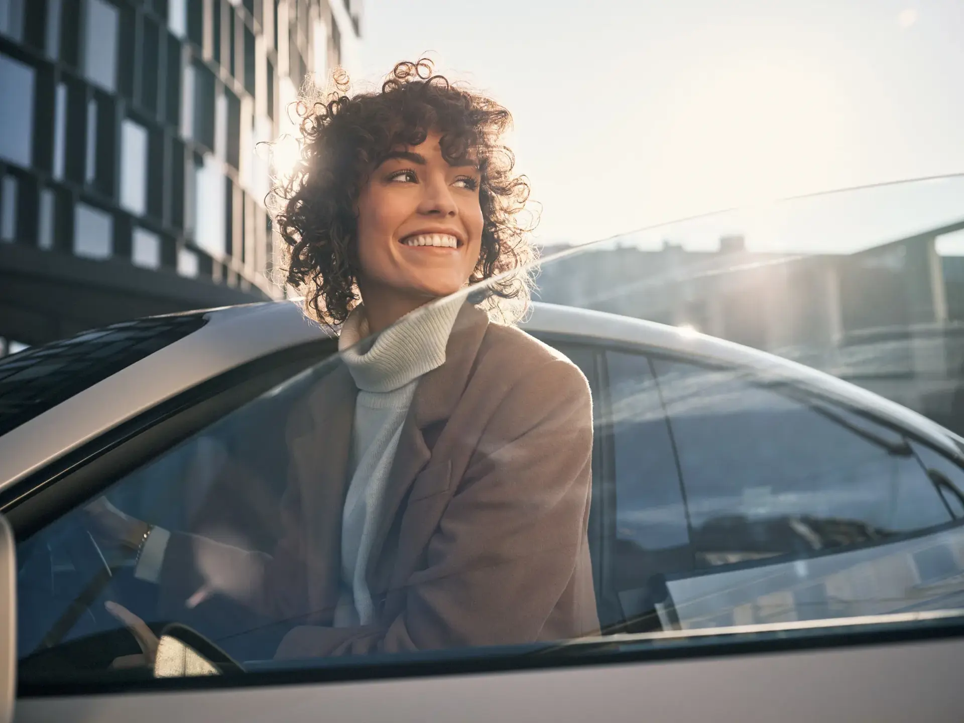 Smilende person i elegant jakke sidder bag rattet i en lys Porsche i sollys foran en moderne bygning.