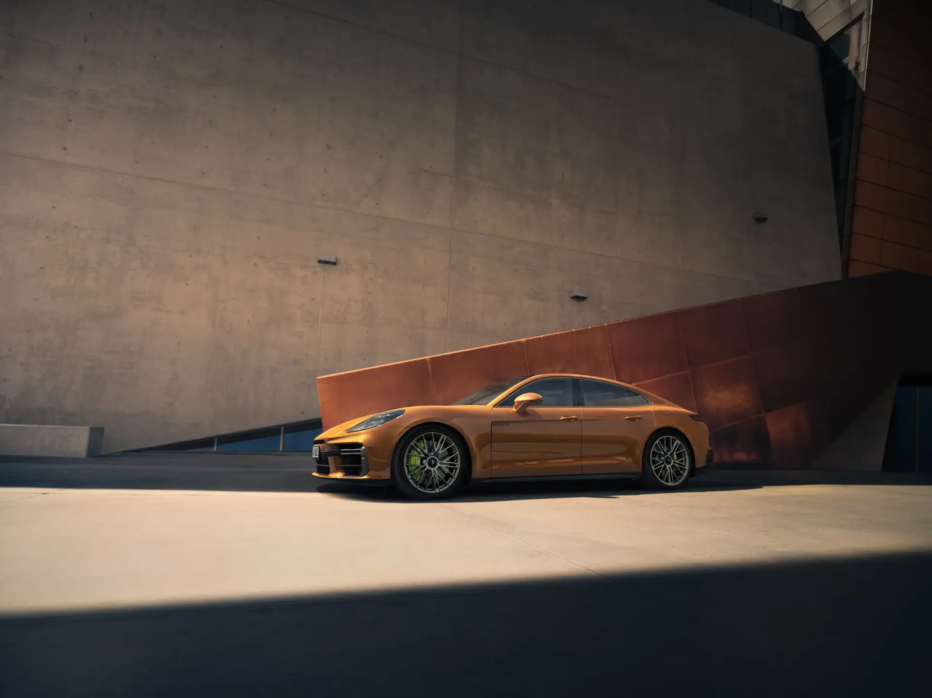 En gyldenbrun Porsche Panamera Turbo E-Hybrid holder parkeret foran en moderne, minimalistisk beton- og stålfacade i skarpt sollys.