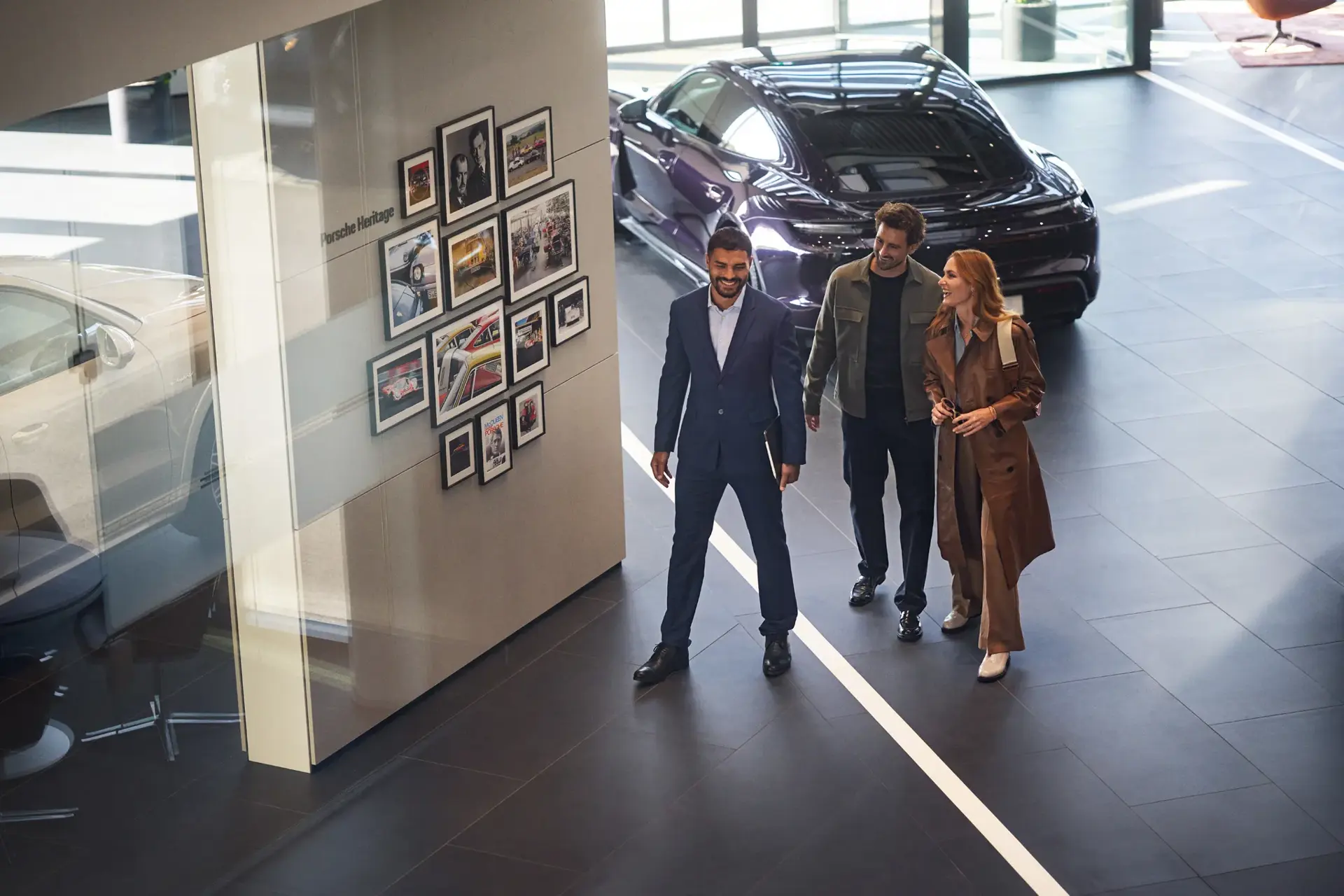 Tre personer går gennem et moderne Porsche-showroom forbi en væg med historiske billeder og en sort sportsvogn.