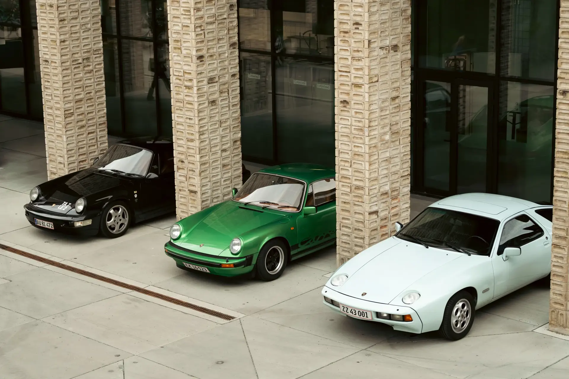 Tre klassiske Porsche-sportsvogne i sort, grøn og hvid holder parkeret mellem søjler foran en moderne bygning.