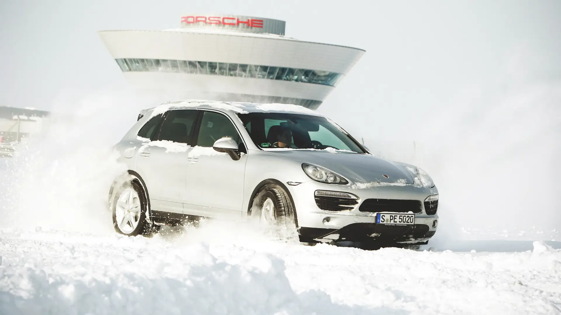 Sølvfarvet Porsche SUV kører gennem sne foran et moderne Porsche-bygning i baggrunden.