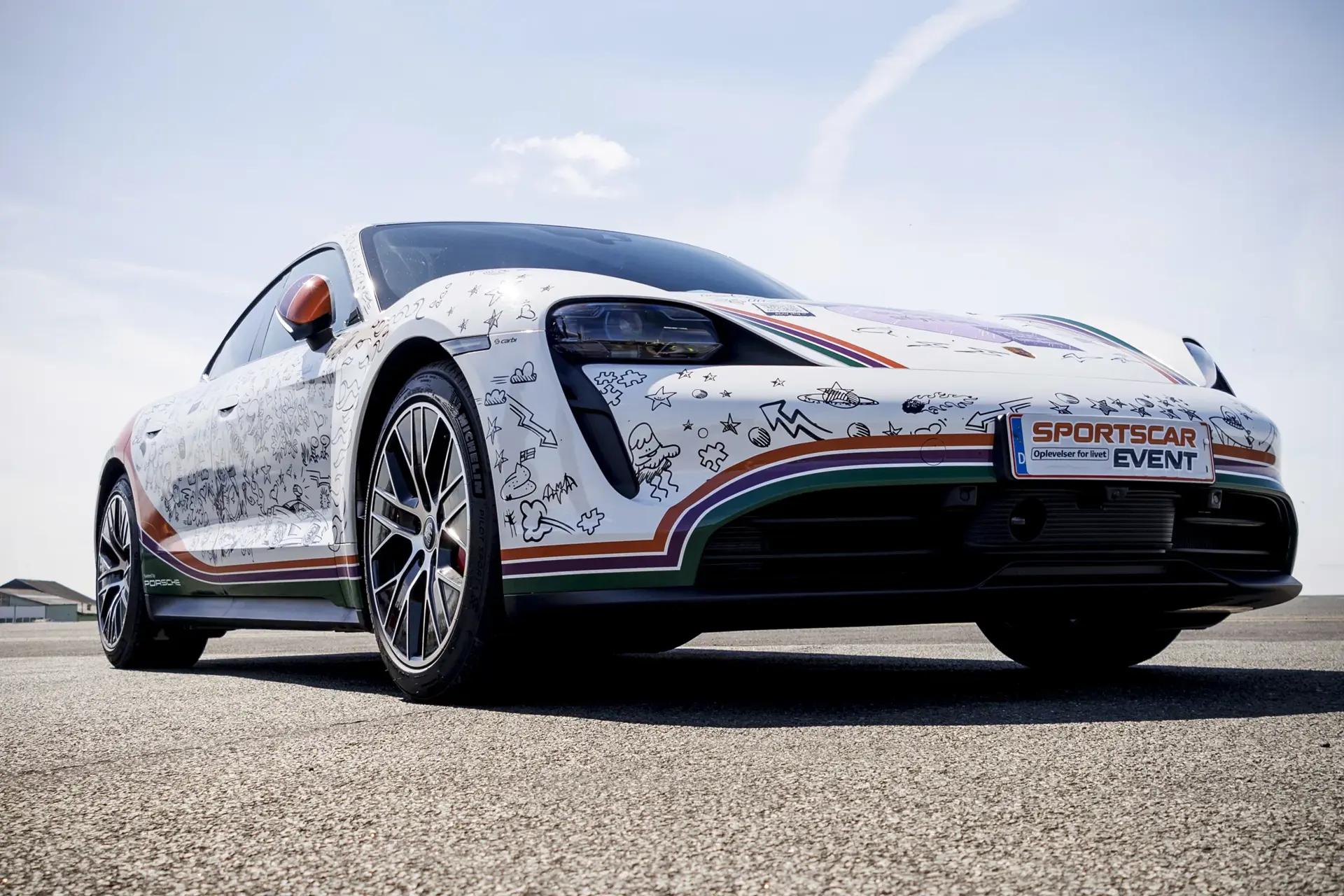En hvid Porsche sportsvogn med farverig, tegnet dekorationsfolie holder på en racerbane under et klart blå himmel ved et Sportscar Event.