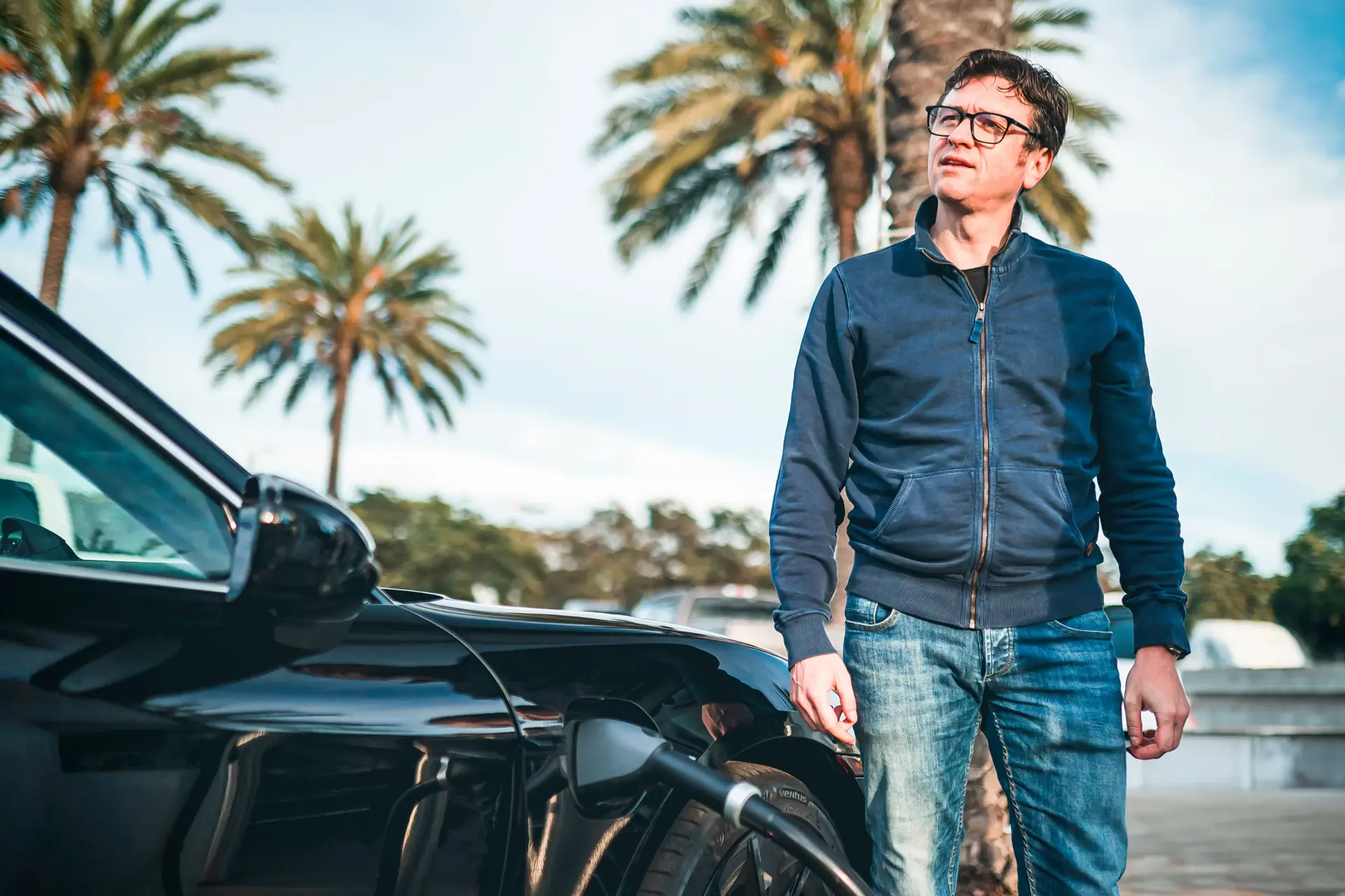 Mand står ved en sort Porsche Taycan, der er tilsluttet en ladestander foran palmer i baggrunden.