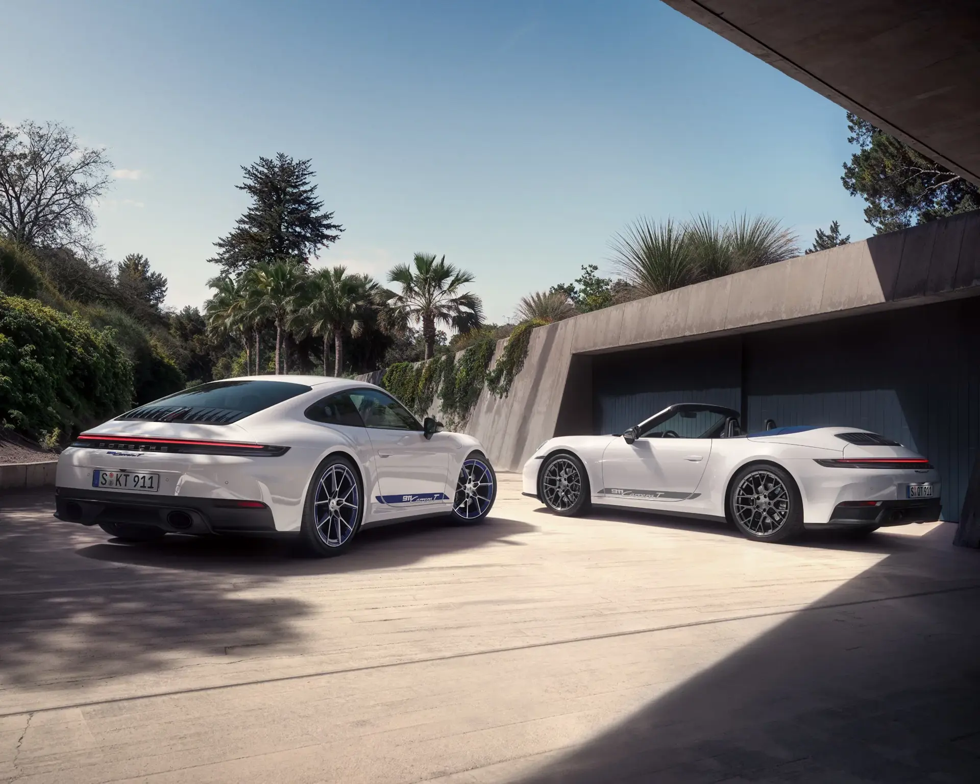 To hvide Porsche 911 Carrera T sportsvogne, én coupé og én cabriolet, holder parkeret i en moderne indkørsel omgivet af palmer.