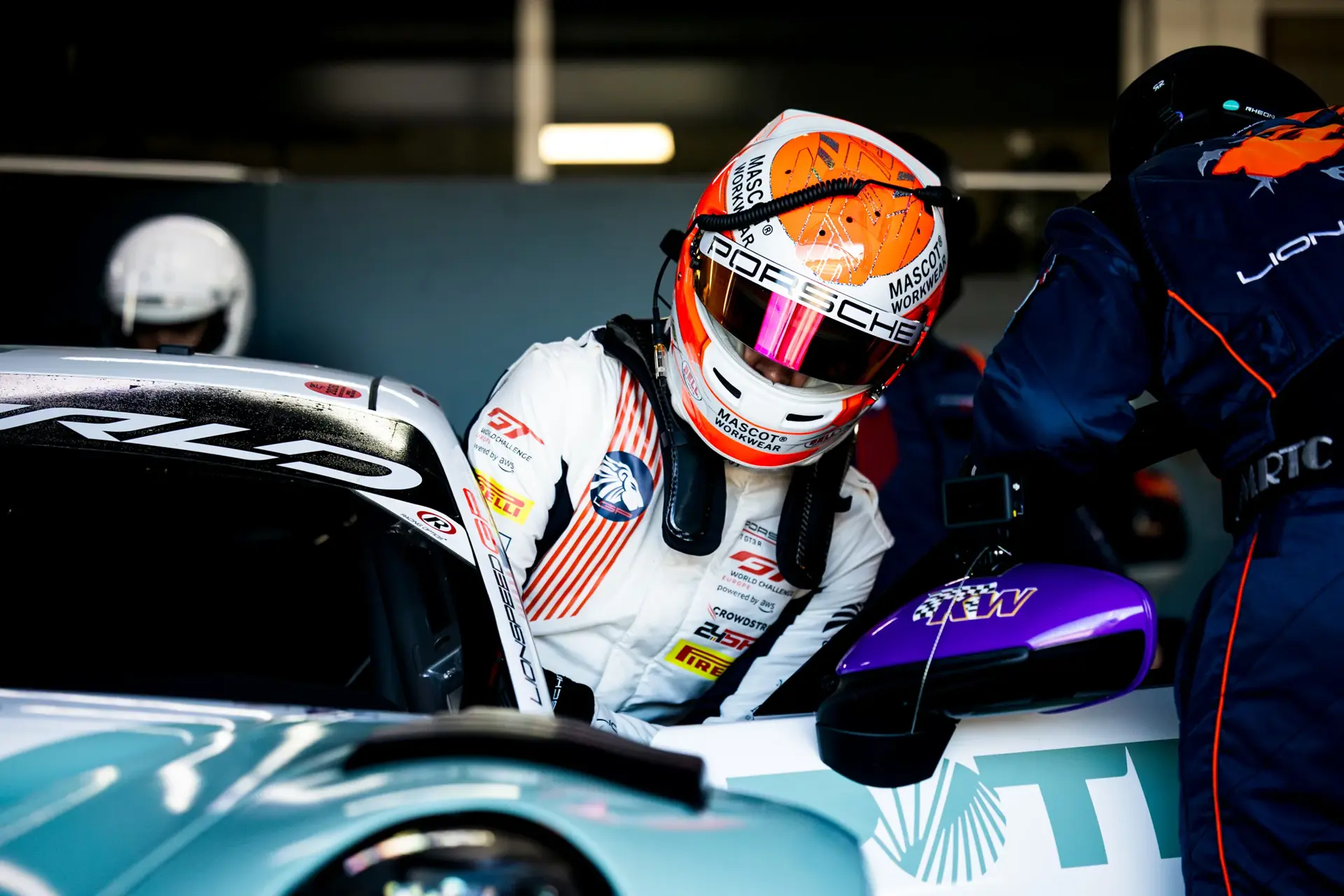 Racerkører i hvid dragt og orange hjelm stiger ned i en Porsche-racerbil i pitten under et løb.