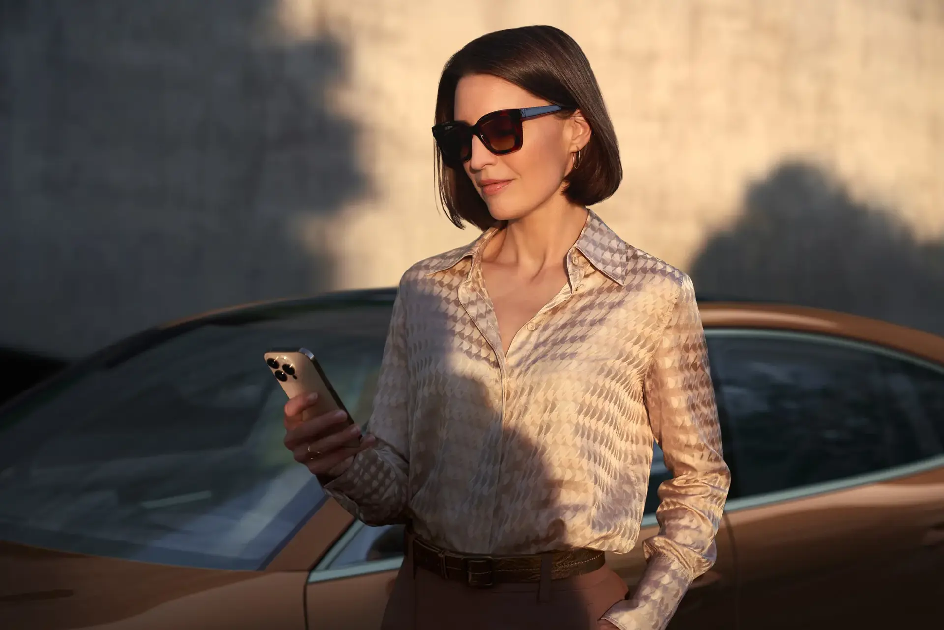 Kvinde med solbriller står foran en brun Porsche Panamera og ser på sin smartphone.