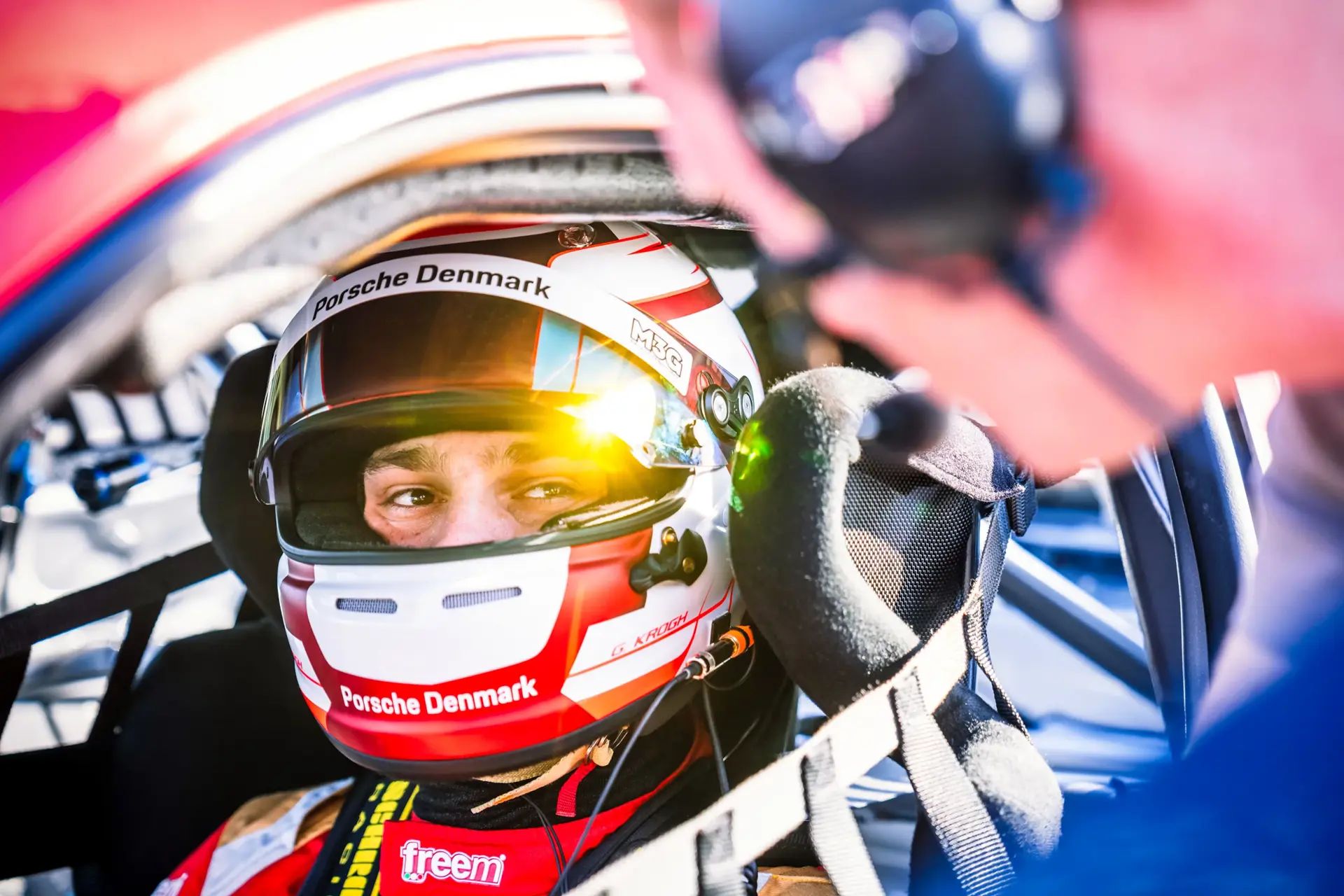 Racerkører med Porsche Denmark-hjelm sidder spændt fast i cockpittet og taler med en person uden for bilen før løbet.