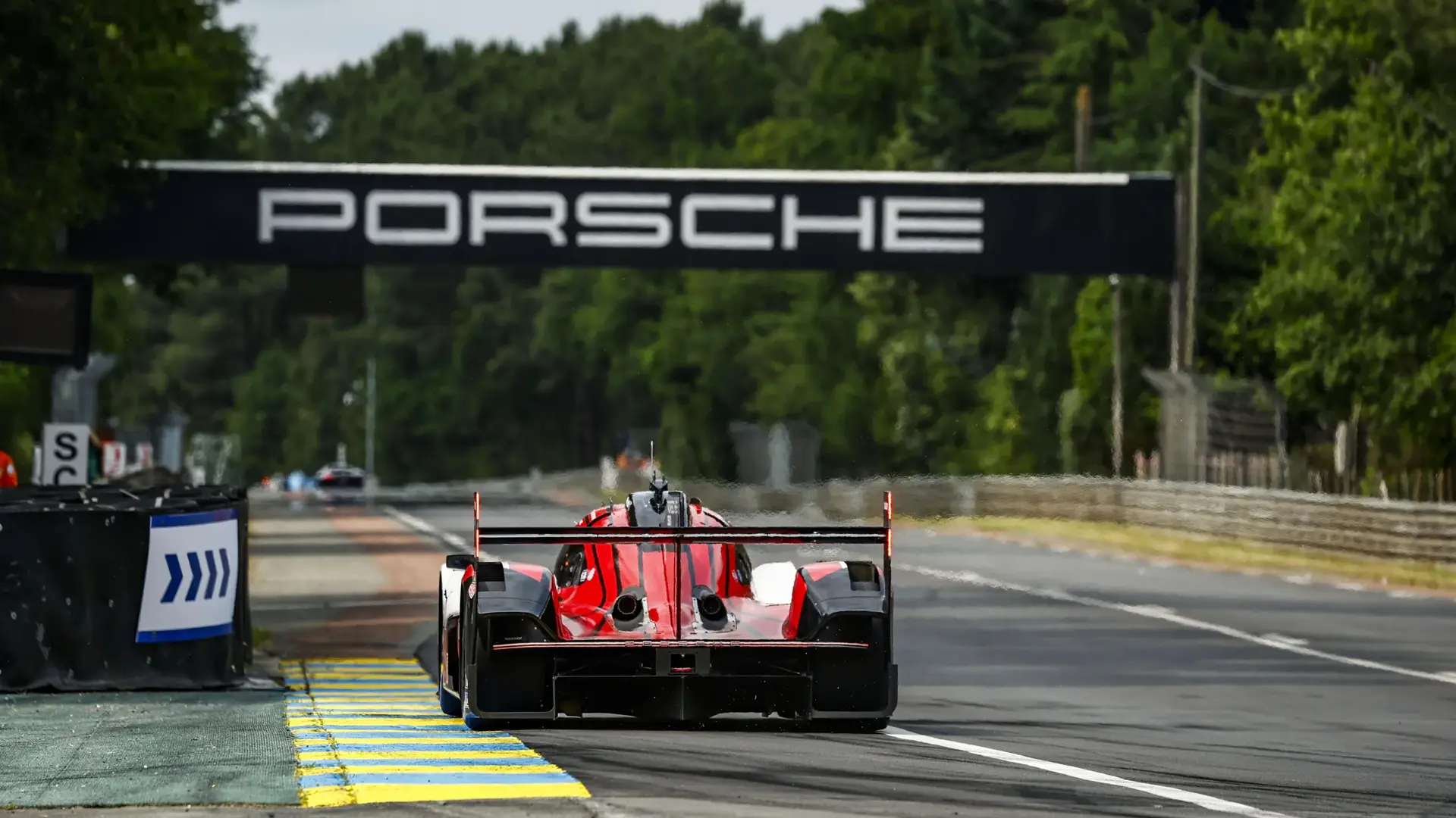 En rød Porsche LMP-racer kører ned ad langsiden på Le Mans-banen under en sort Porsche-bro.